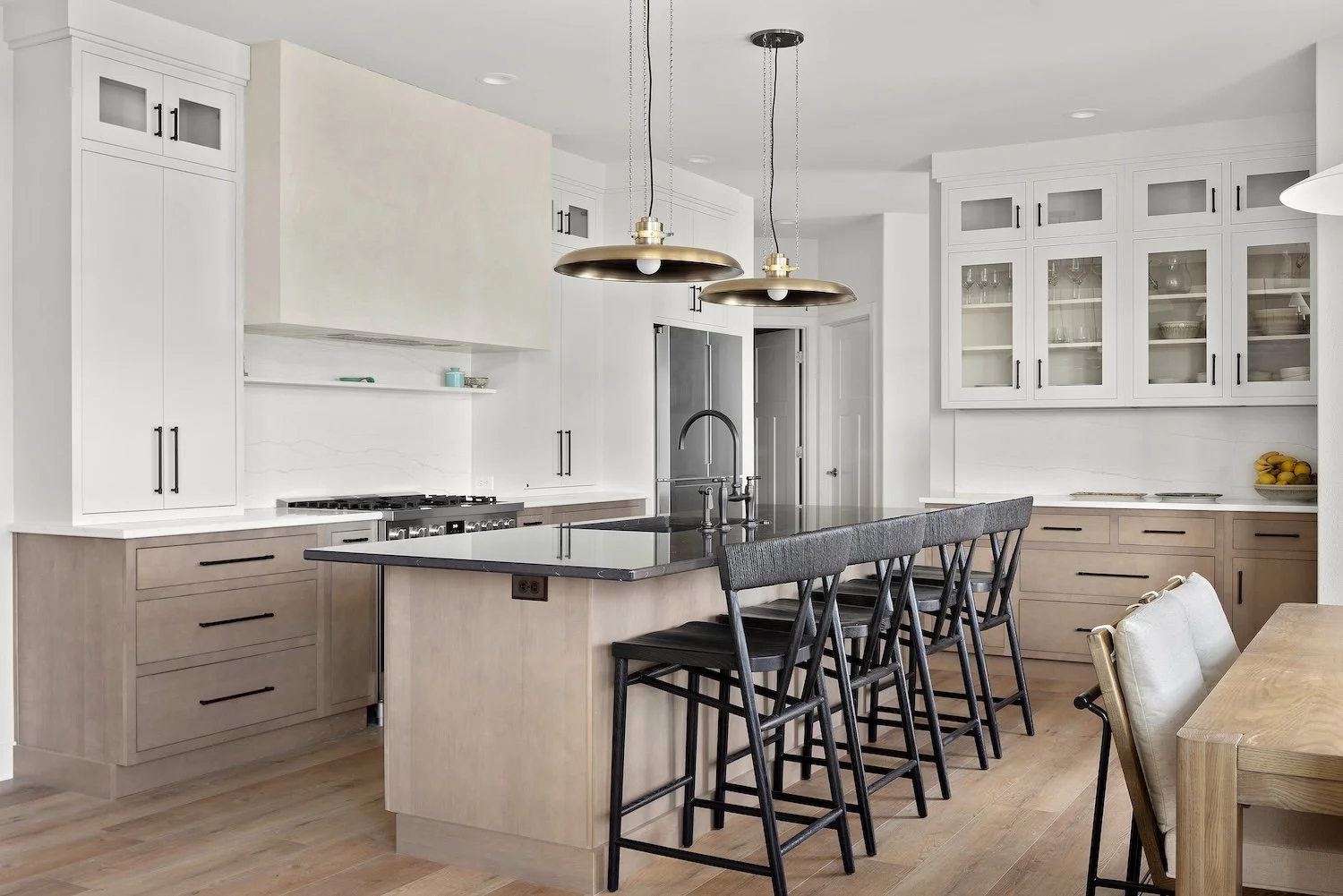 Modern kitchen with light wood cabinets, white upper cabinets with glass doors, black handles, a black countertop island with a sink, black bar stools, and gold pendant lights