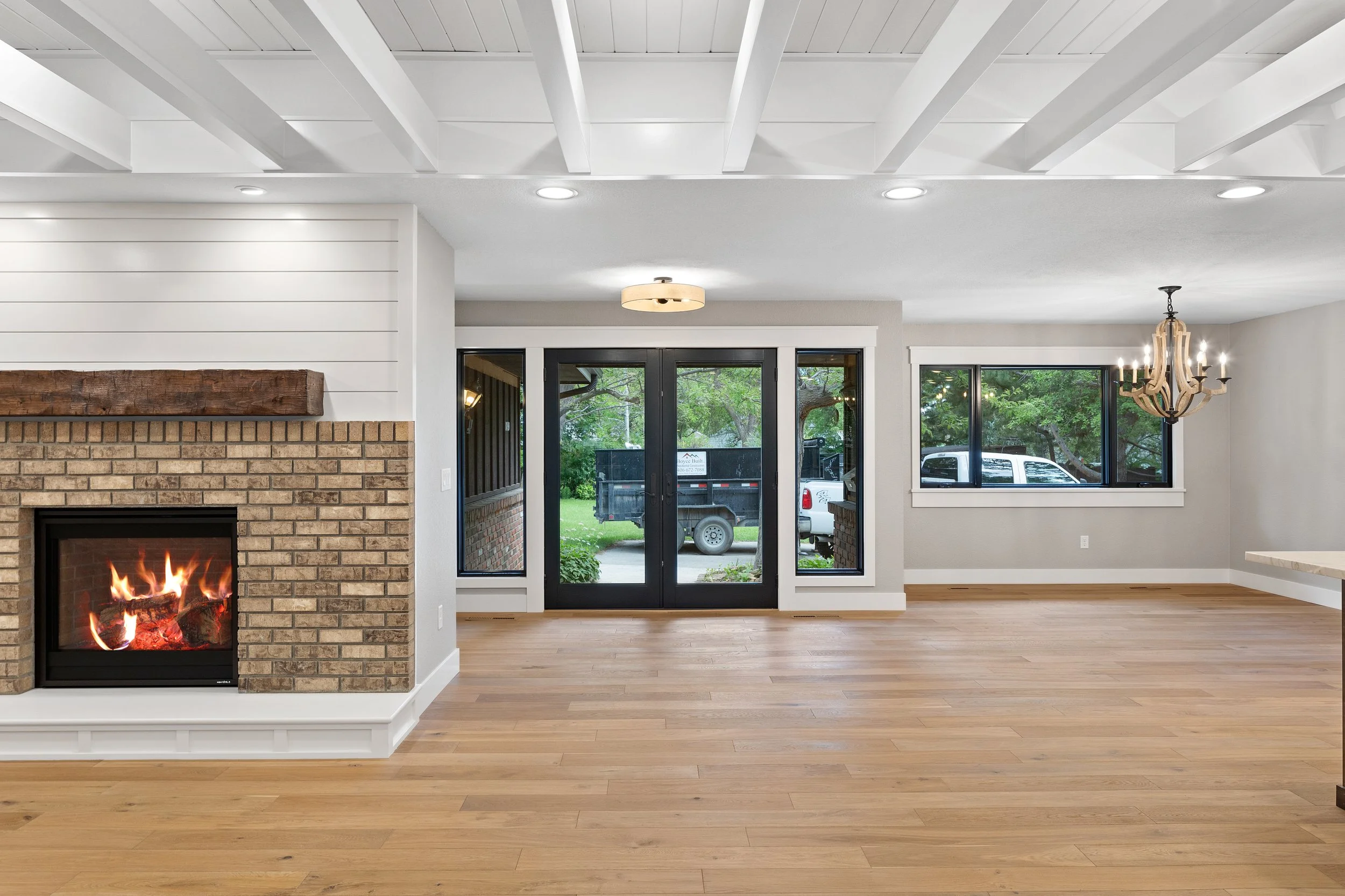 Empty living room with a burning fireplace, wooden floor, multiple windows, sliding door leading outside, and a chandelier.