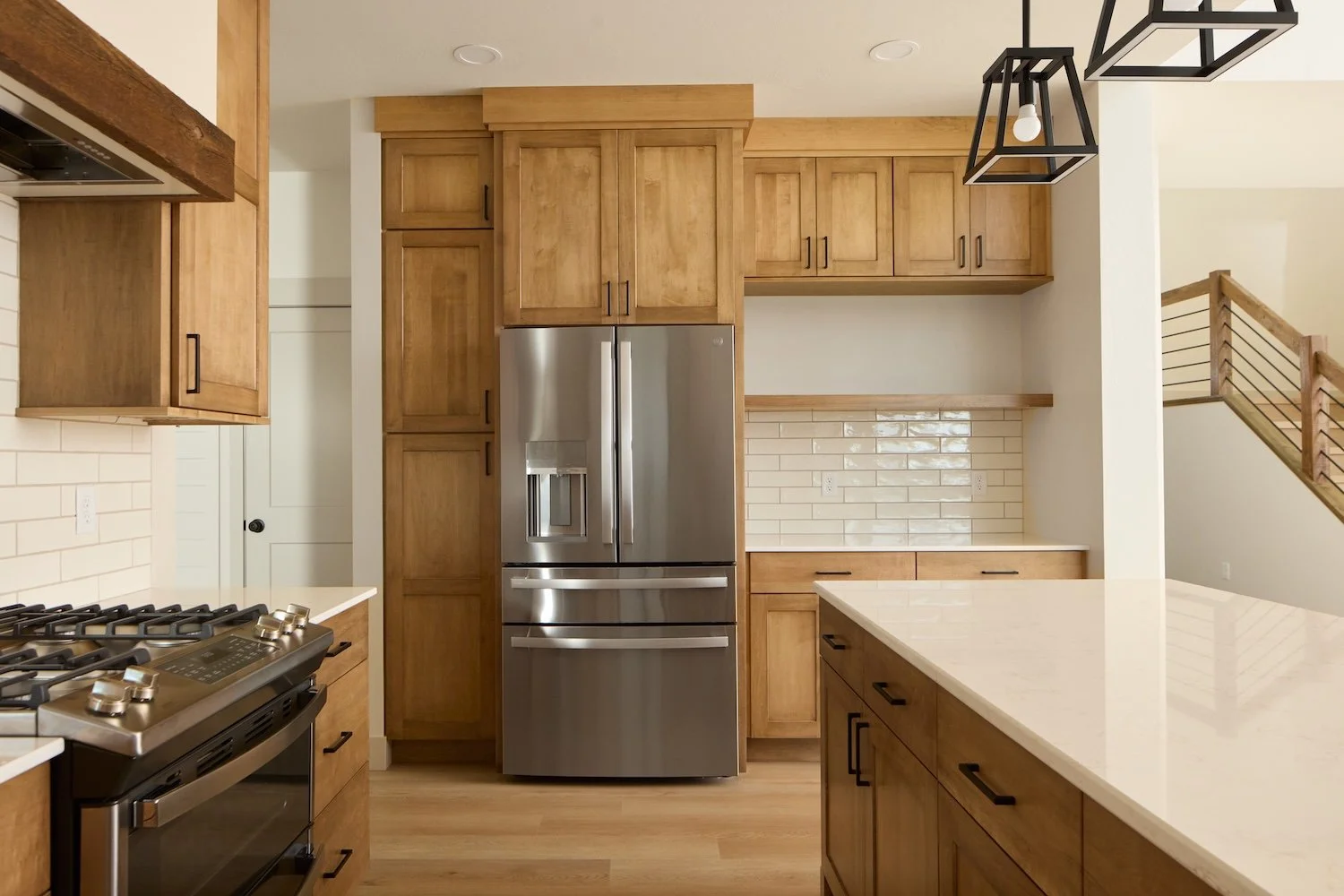 Modern kitchen with wooden cabinets, stainless steel refrigerator, white countertops, brick backsplash, and a stove. Open space with staircase nearby.