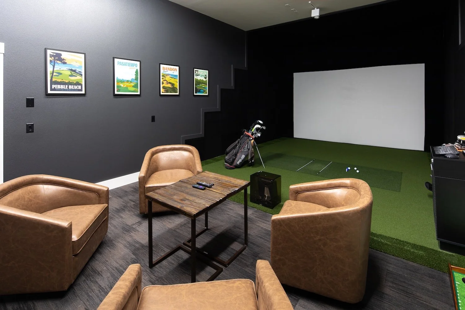 Indoor golf simulation room with a small green artificial turf area, golf clubs in a stand, a white screen, and a camouflaged leather armchair set with a wooden table and remote controls in the foreground. Three colorful posters on the dark wall depict golf courses.