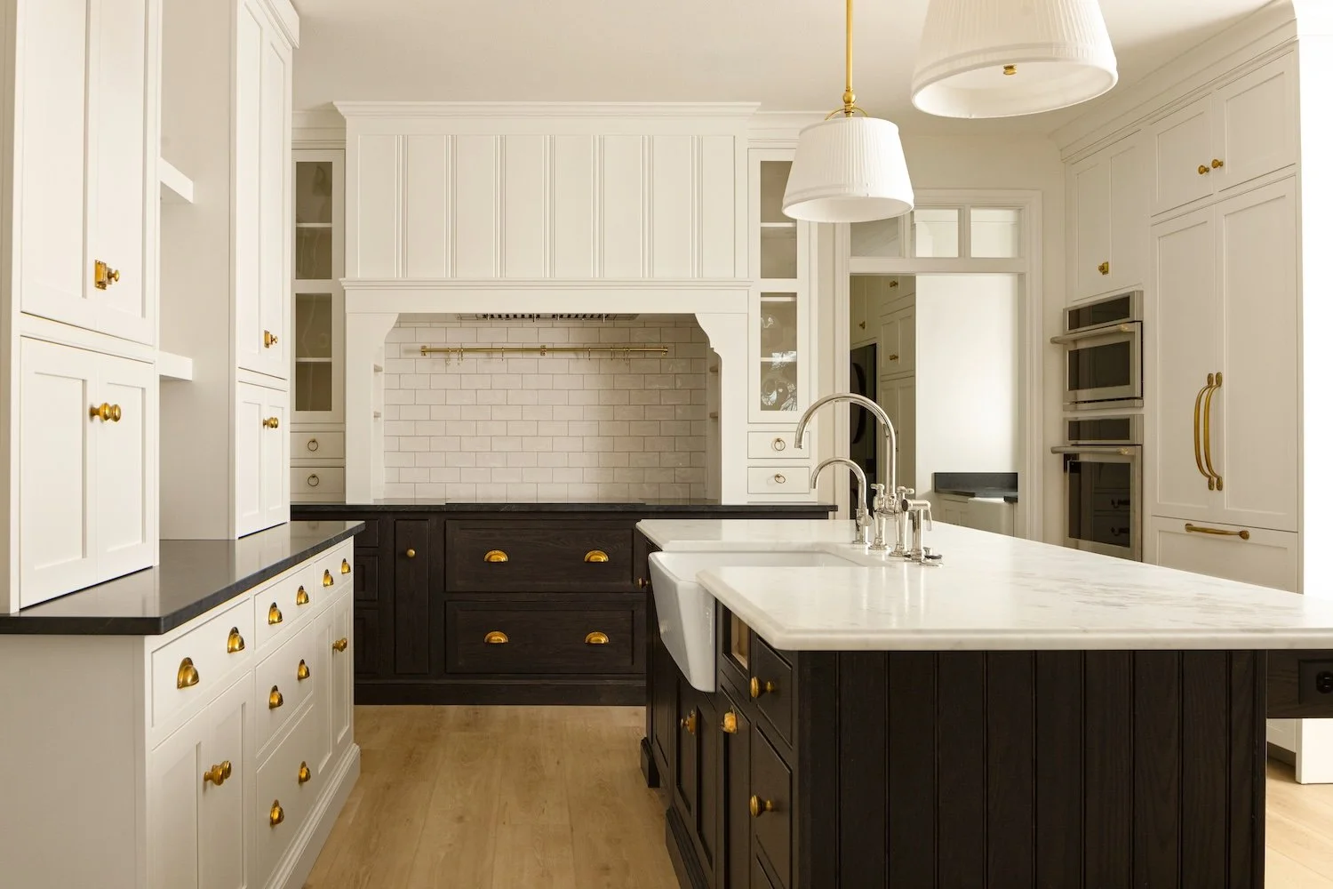 Modern kitchen with white cabinetry, black island with gold hardware, white marble countertops, and a white subway tile backsplash. Overhead lighting fixtures hang above the island.