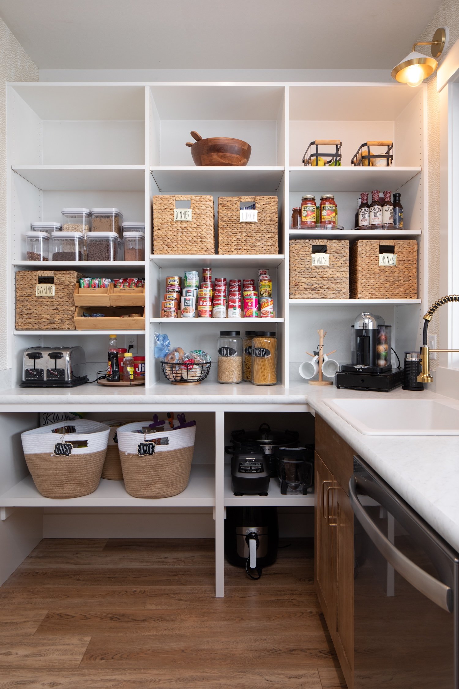Kitchen pantry with white shelves holding baskets labeled for baking, snacks, breakfast, drinks, and containers of cereals and canned goods. Below are storage baskets labeled for chips and snacks, with small kitchen appliances and a trash can on the floor. The counter has a coffee maker and a sink with a gold faucet.