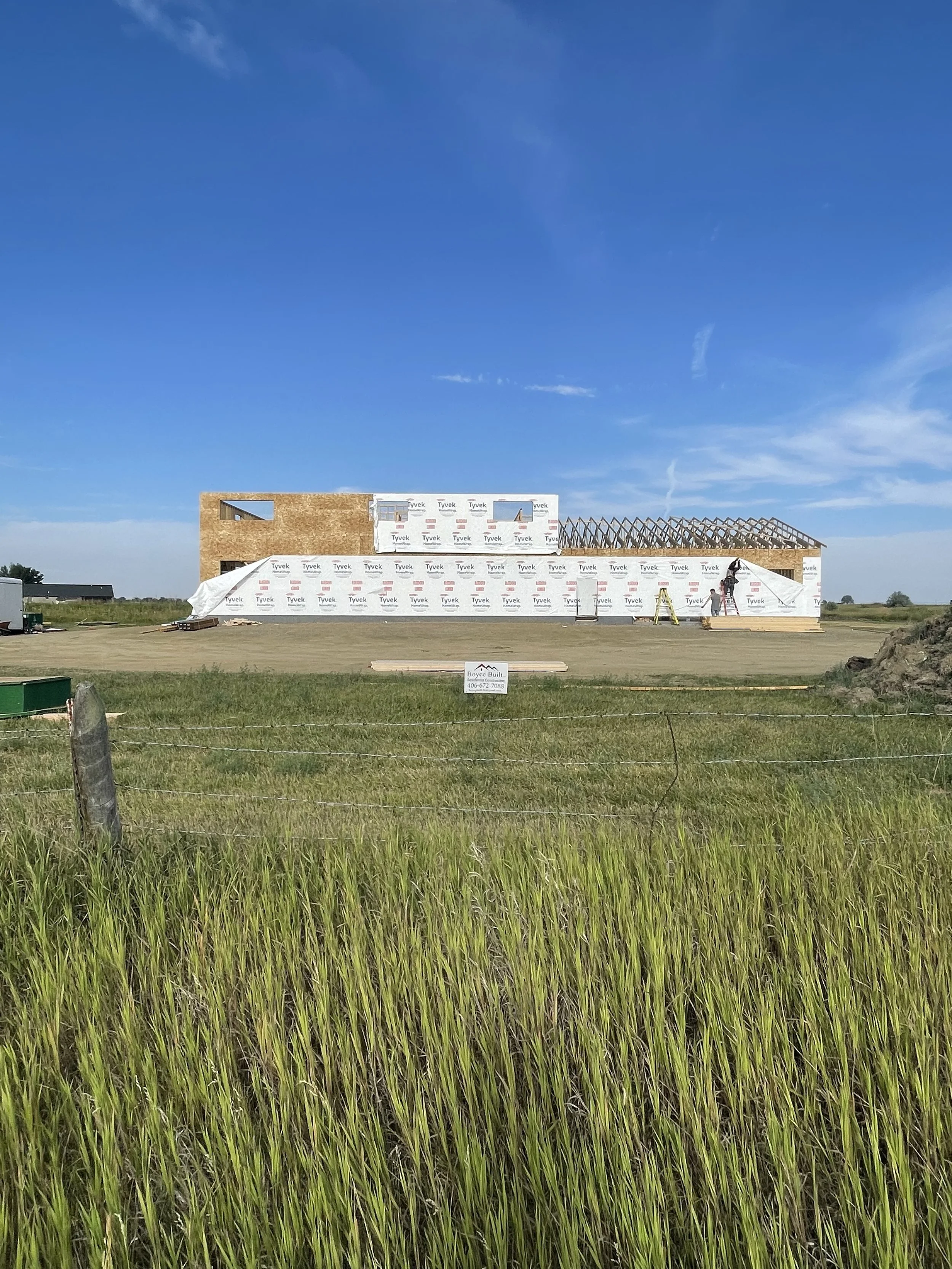 Construction site of a house with partially built wooden framing and wall sheathing, surrounded by green grass and a barbed wire fence, under a Montana clear blue sky. General Contractor sign- Boyce Built Construction