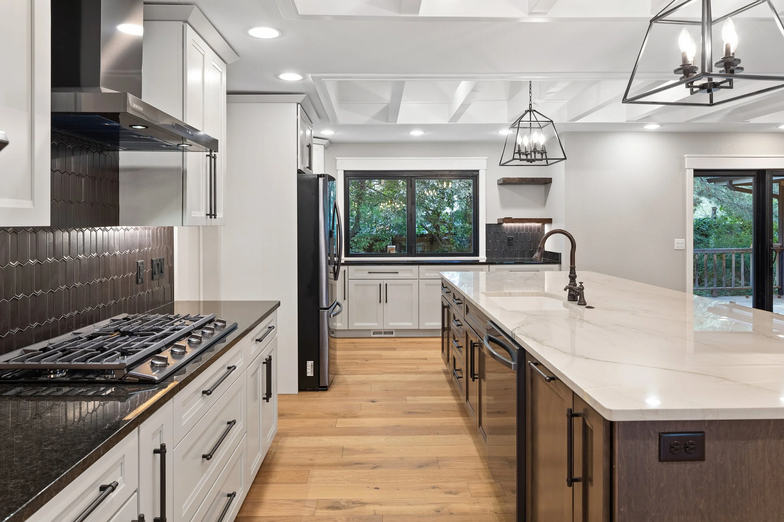 Modern kitchen with white cabinets, a large center island with a marble countertop, black fixtures, wooden flooring, and large windows showing greenery outside.