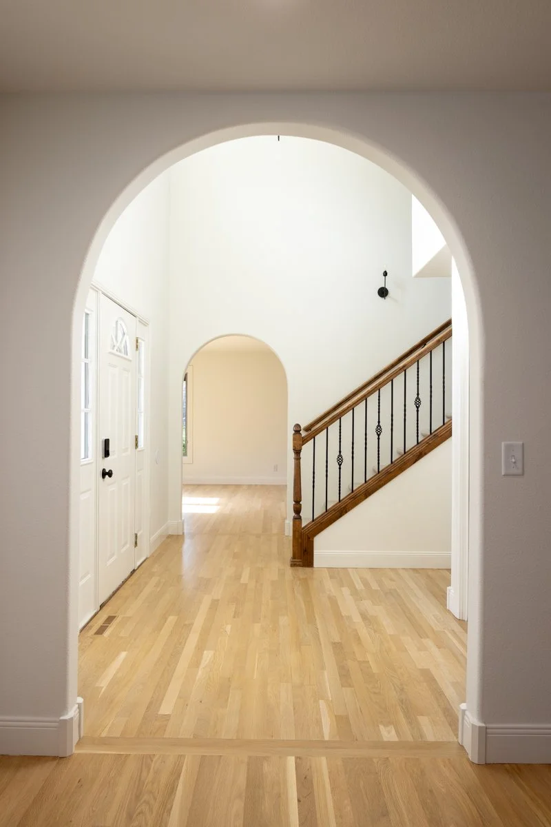 Interior view of a house entryway with a front door, hardwood flooring, and staircase with wooden handrail and black wrought iron spindles, seen through an arched doorway.