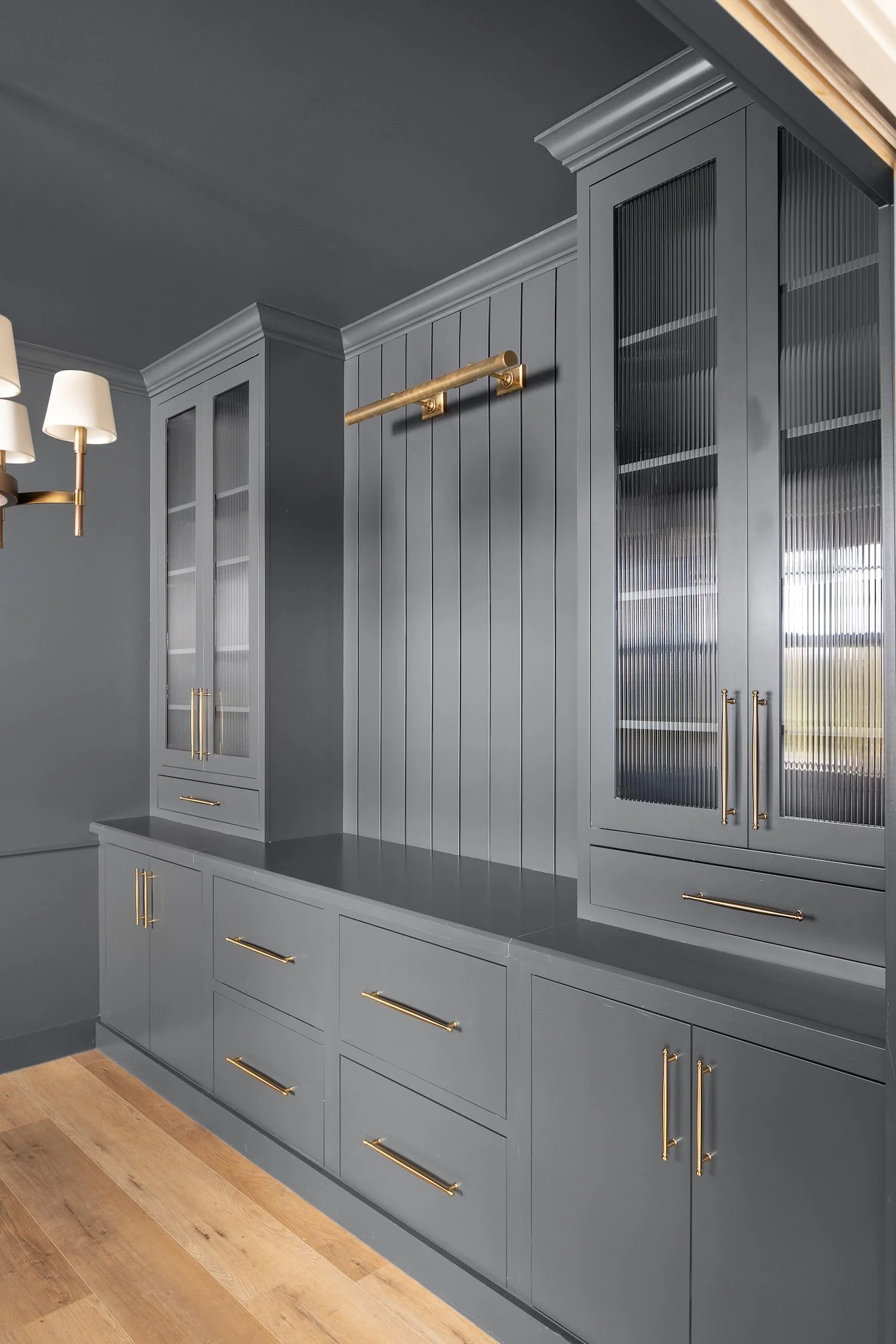 A kitchen with gray cabinets and gold hardware, a brass rail, and a wooden floor. The kitchen includes a built-in cabinet with glass doors and vertical ribbing, a countertop, and a wall-mounted brass light fixture.