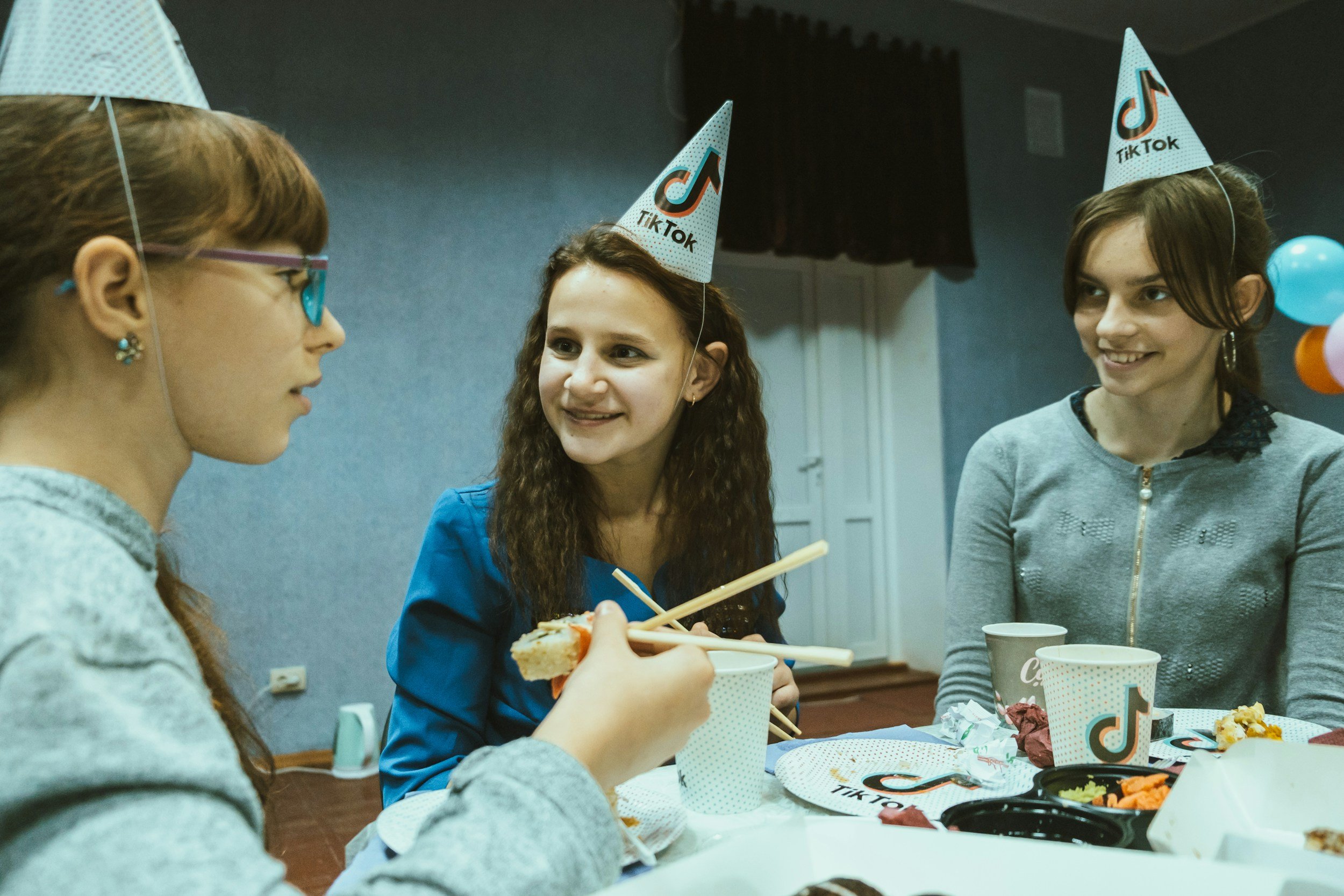 Three young women wearing TikTok party hats are sitting at a table with food and drinks, smiling and engaging in conversation.