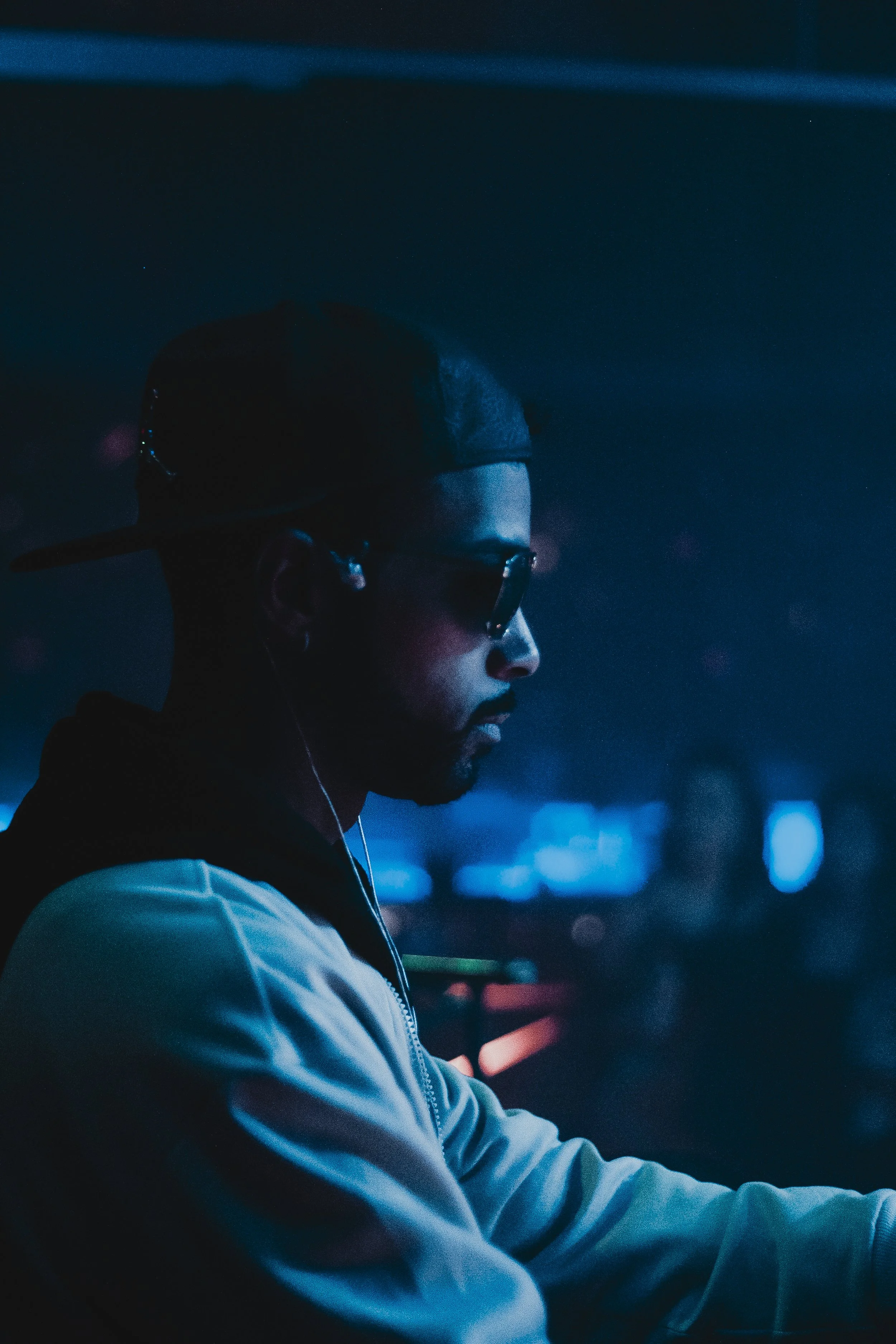 Profile of a young man wearing glasses, a backwards baseball cap, and earphones, illuminated by city lights at night.