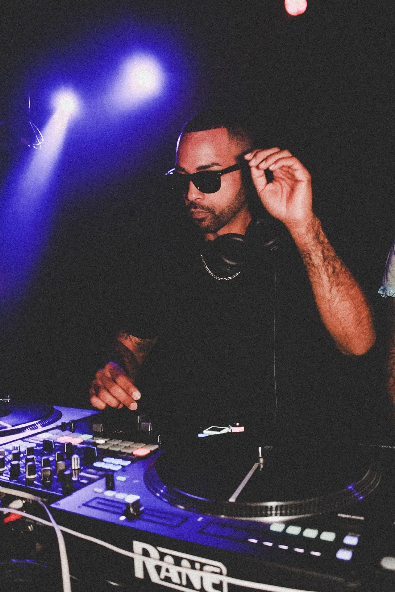 A male DJ wearing sunglasses and headphones around his neck adjusts his glasses while working at a DJ booth with colorful controls, illuminated by purple and blue lighting.