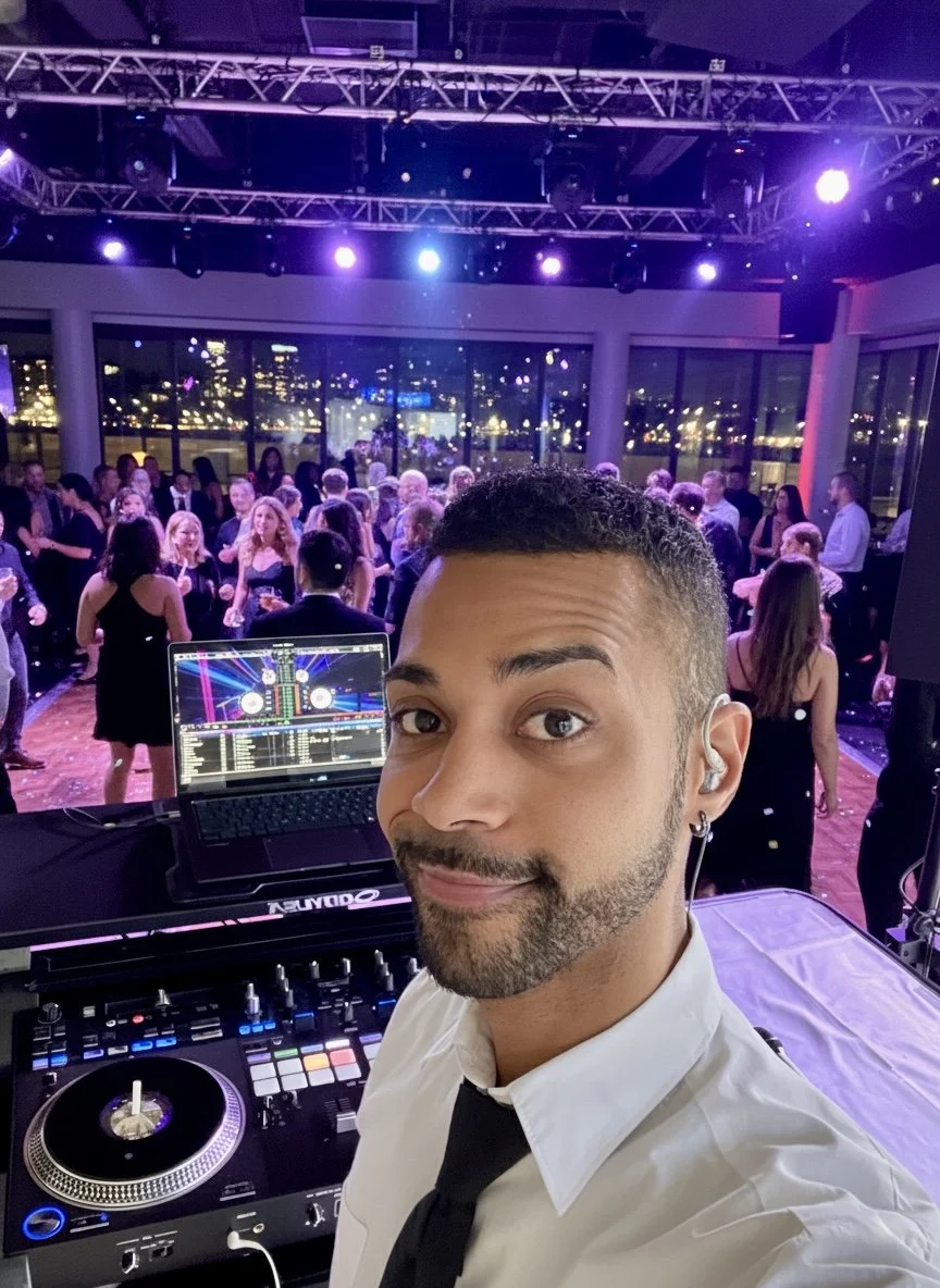 A DJ taking a selfie at a crowded dance party with many people dancing and socializing, city lights visible through the large windows.