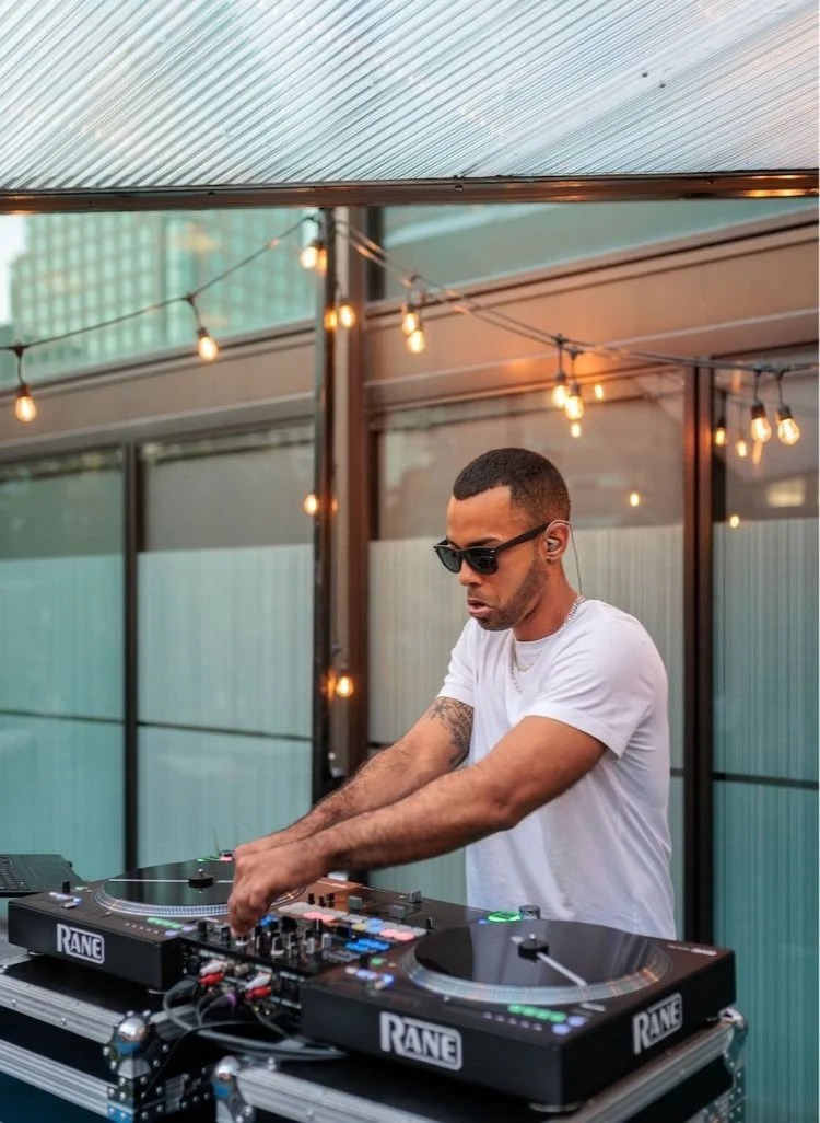 A male DJ wearing sunglasses and a white t-shirt is mixing music with turntables outdoors, under string lights and a glass roof, with modern buildings in the background.