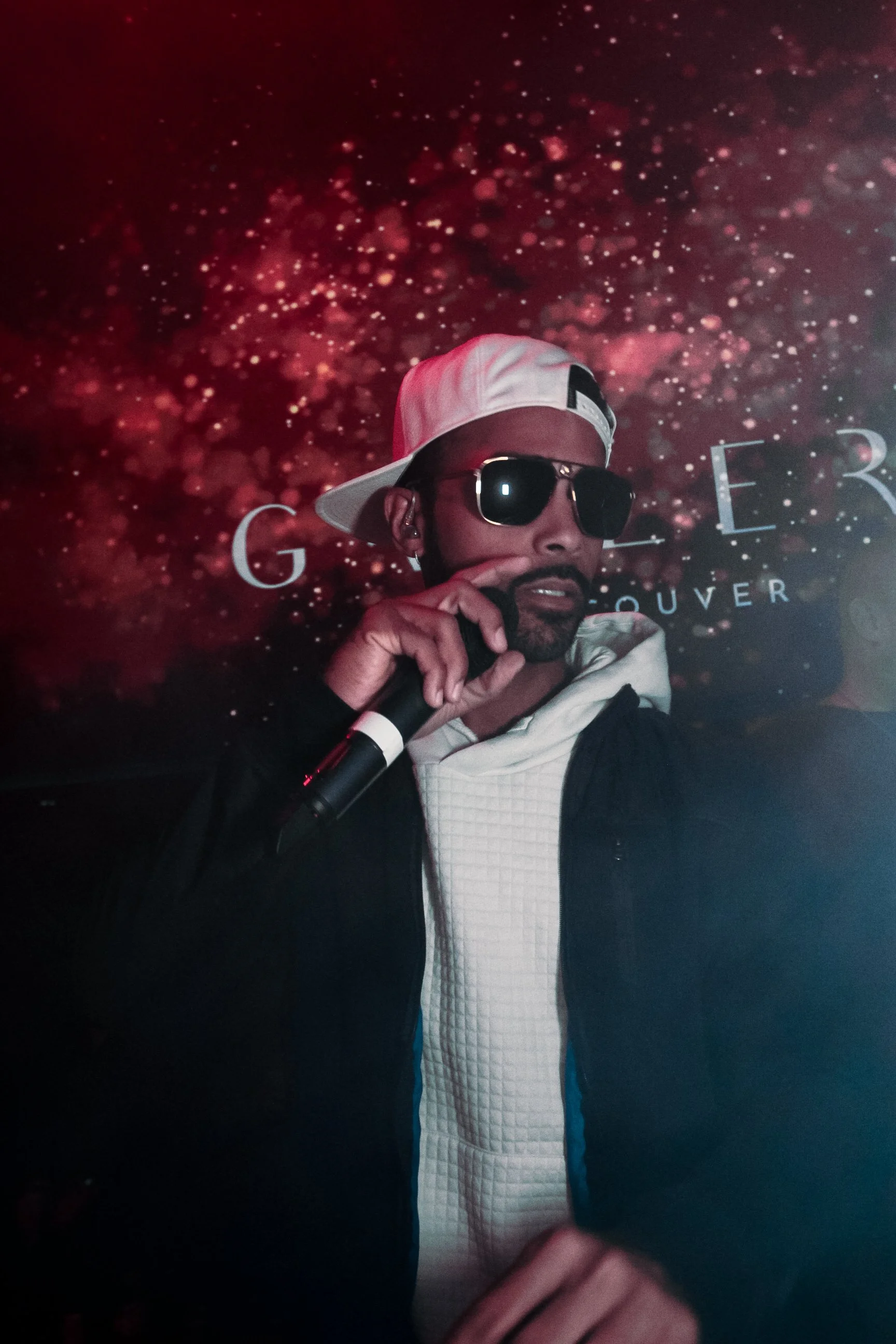 A man with a beard wearing a white cap, sunglasses, a white hoodie, and a black jacket holding a microphone.