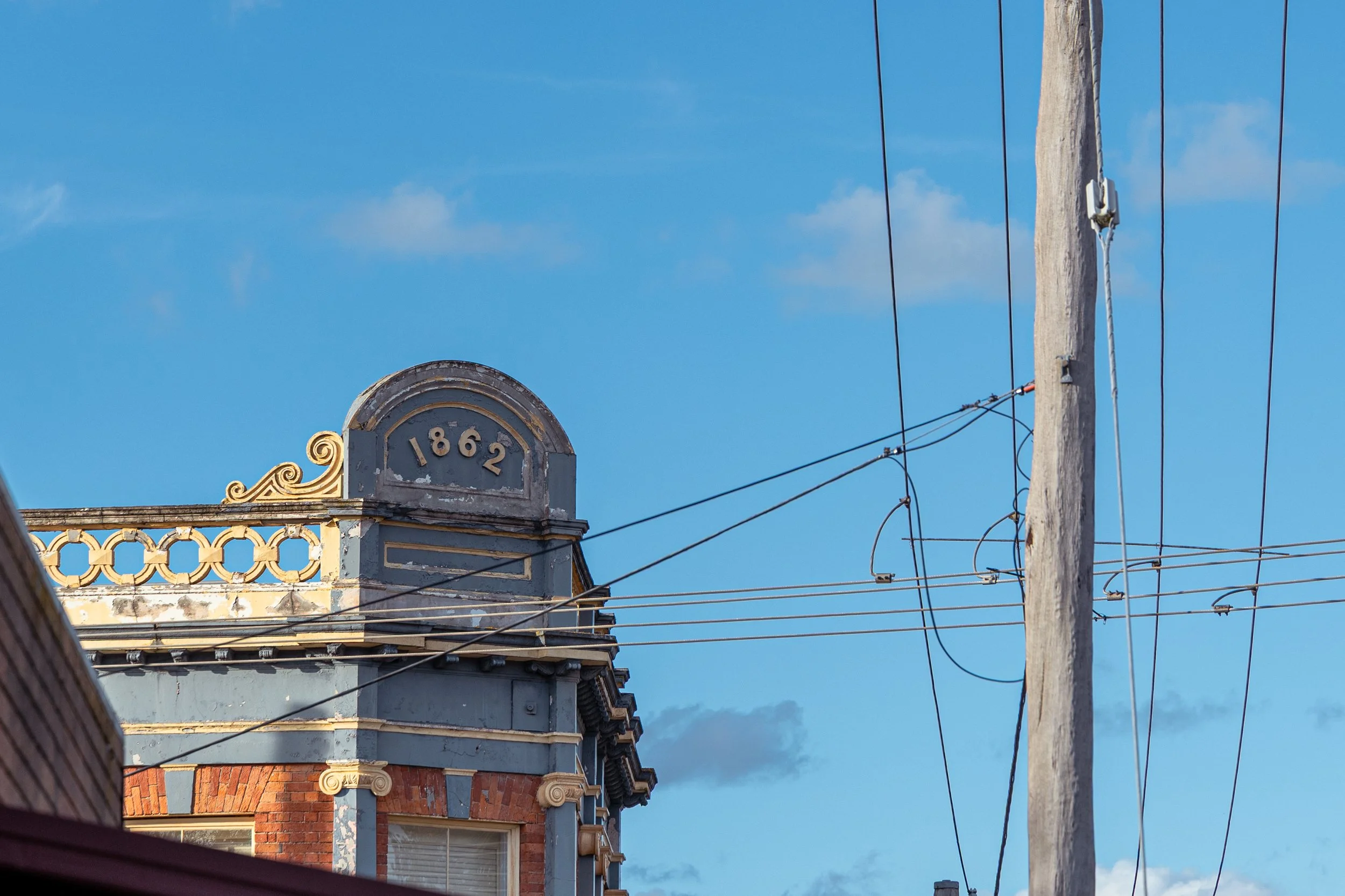 Historic Ballarat building (The Eastern Hotel, built in 1862) with ornate architectural details, a power pole and overhead wires against a bright blue sky.