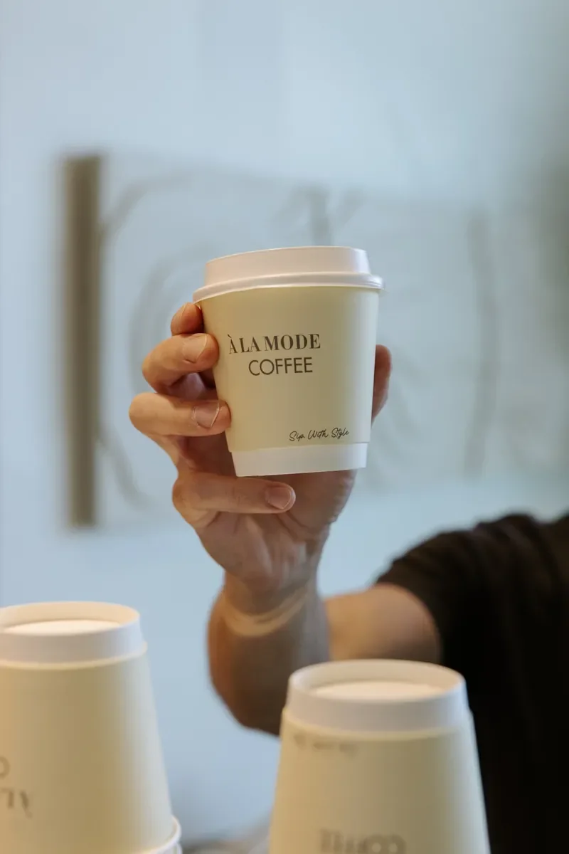 Hand holding a cream À LA MODE COFFEE takeaway cup with a white lid.
