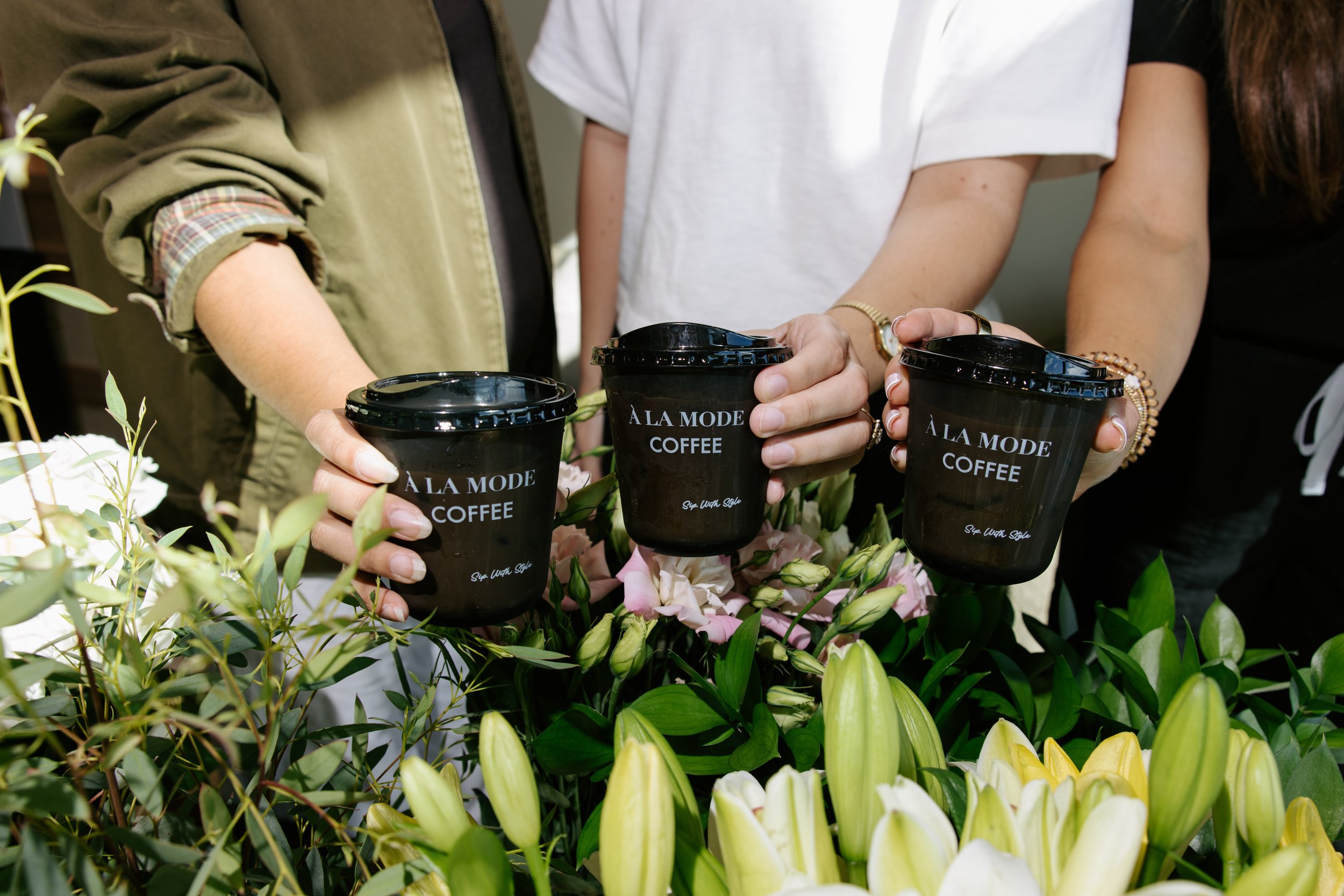 Guests holding black À LA MODE COFFEE cups above a floral display at SkinPharm’s Winter Park grand opening.