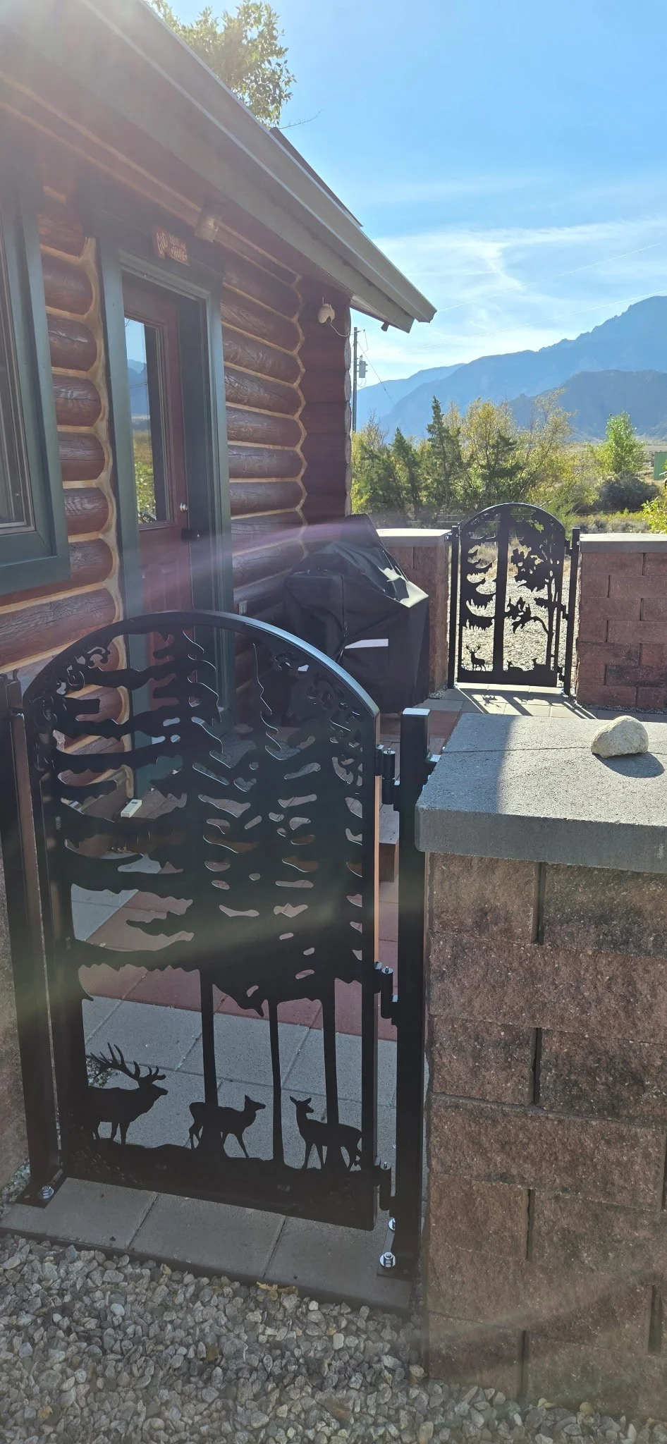 Outdoor patio area with decorative metal gate featuring wildlife silhouette design, stone pillar, and view of mountains and trees in the background.