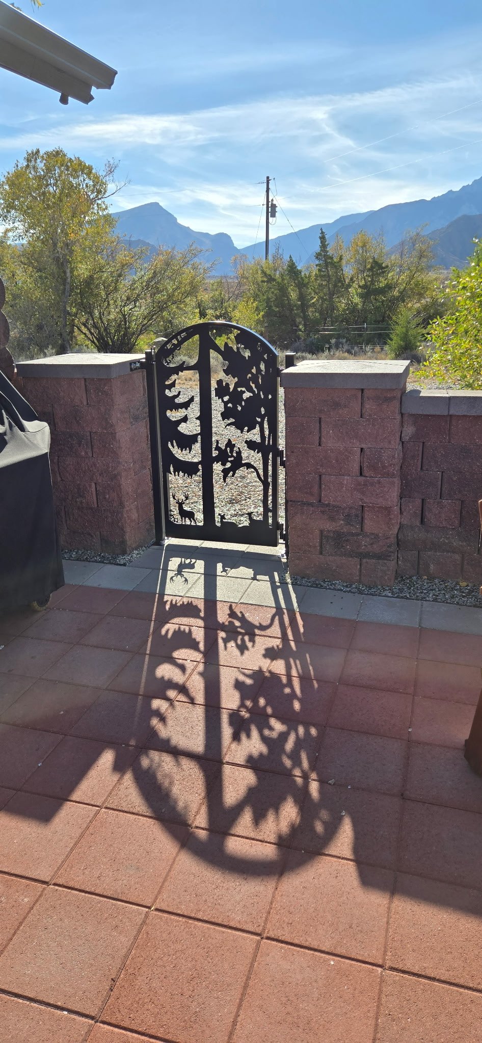View of a decorative metal gate casting a shadow on a tiled patio, with a brick wall, trees, mountains in the background, and clear blue sky.