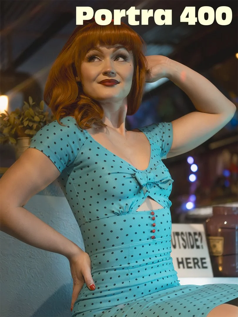 A woman with red hair wearing a blue polka-dot dress with a cutout in the front and red buttons. She is posing with one hand behind her head and looking to the side in a retro-style setting.
