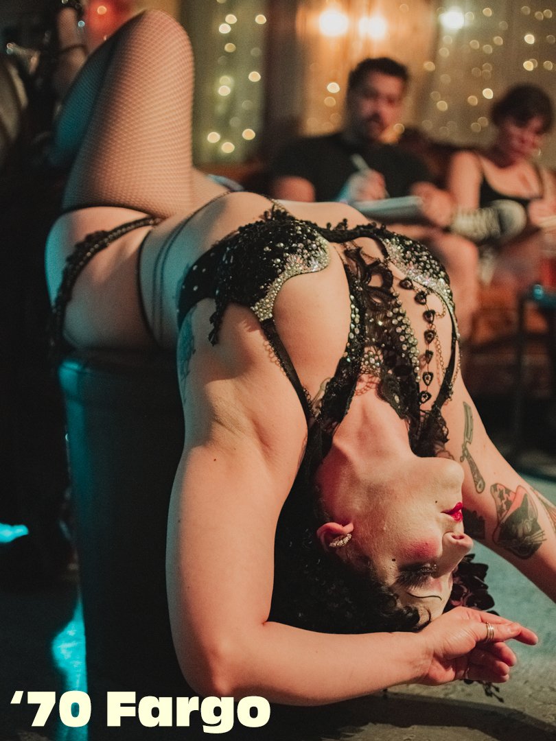 A woman in black lacy and bejeweled lingerie performing a backbend on stage, with two people in the background watching, in a dimly lit setting with string lights.