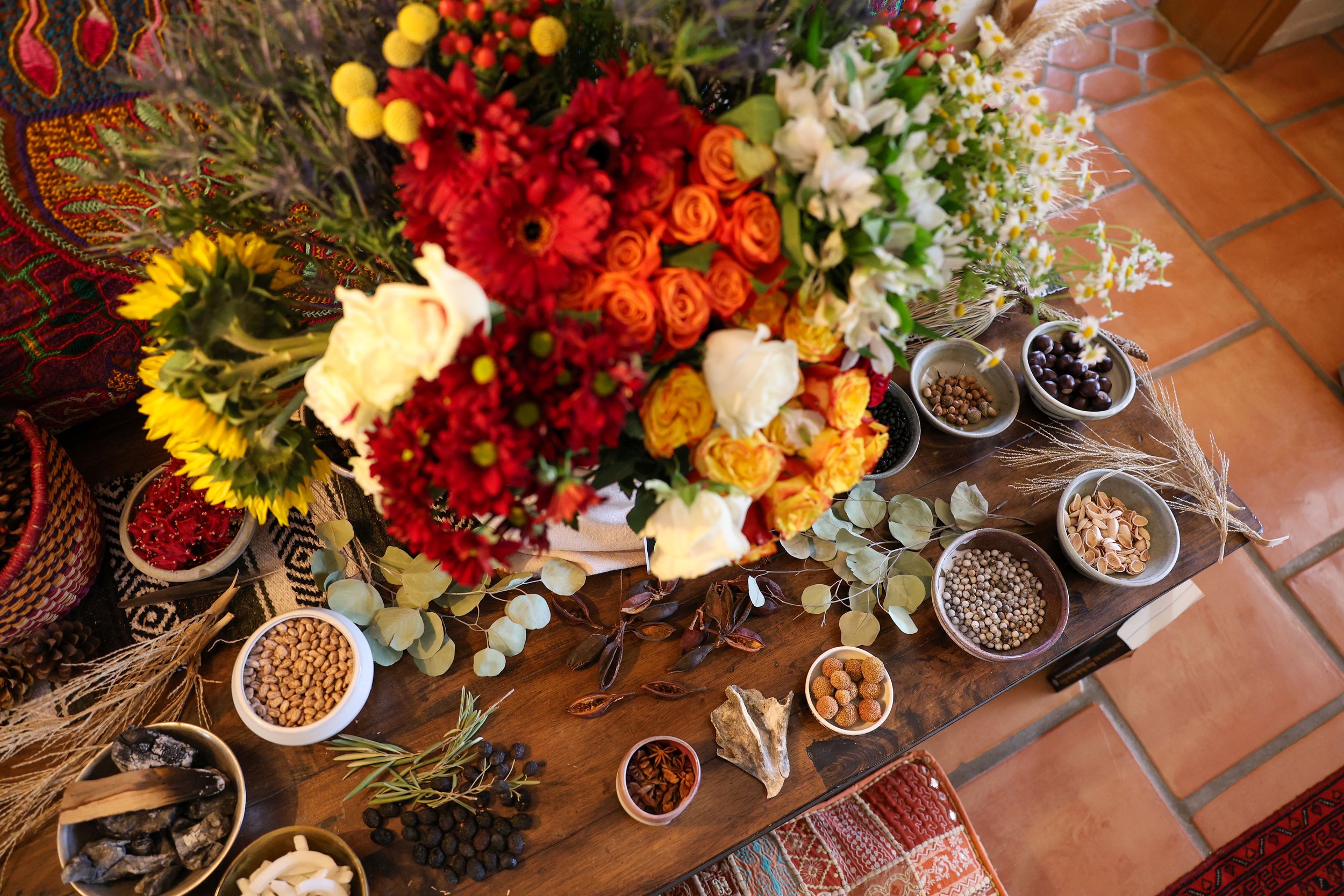 A wooden table with a colorful bouquet of flowers including orange roses, white lilies, and red chrysanthemums, surrounded by various small bowls of herbs, seeds, and natural items. The table is on a terracotta-tiled floor.