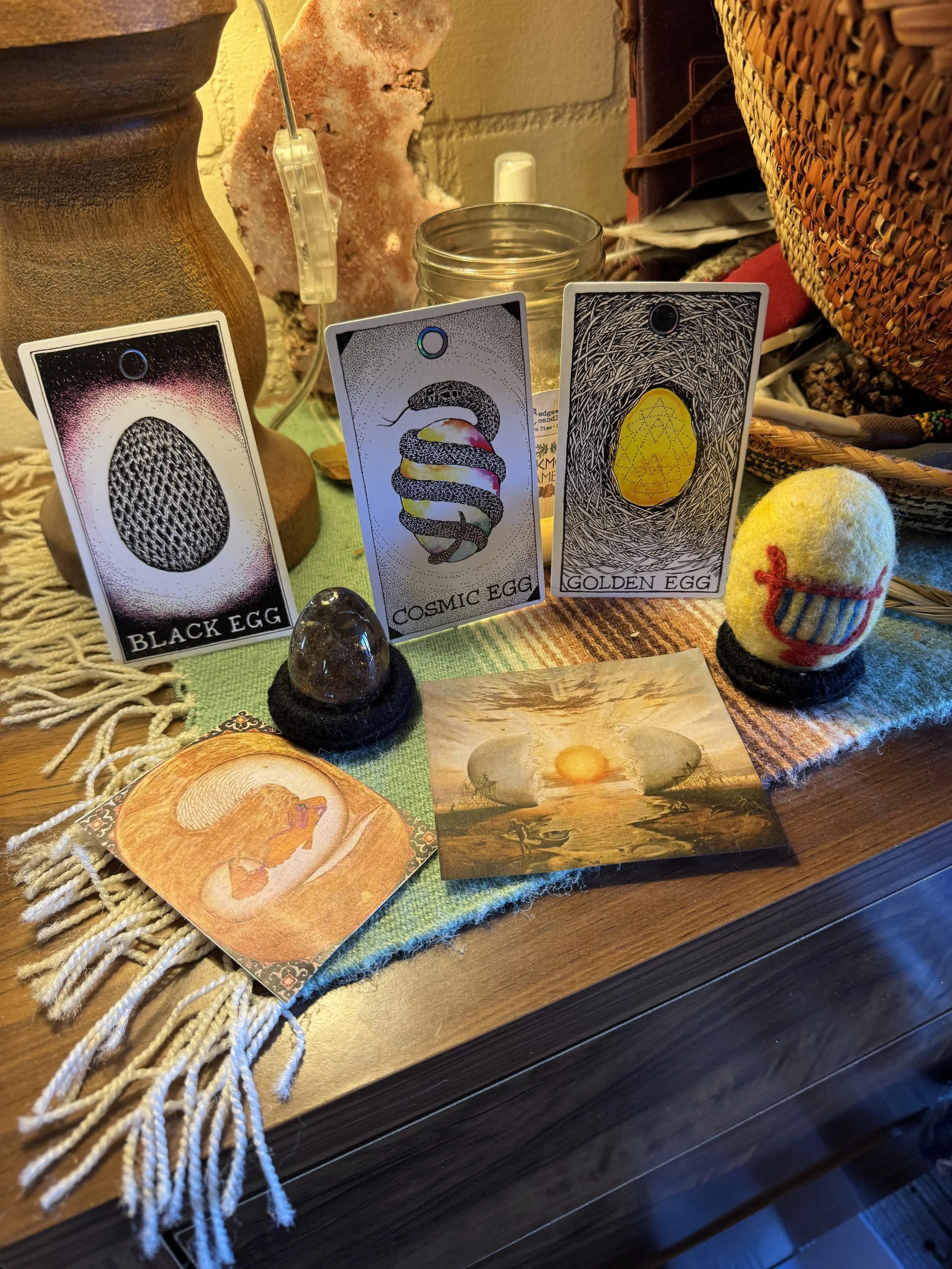 A tabletop display with fortune-telling cards, a small felted egg with a face, a framed picture of a sunrise over a pond, and various decorative objects including a mineral, a basket, and a small jar.