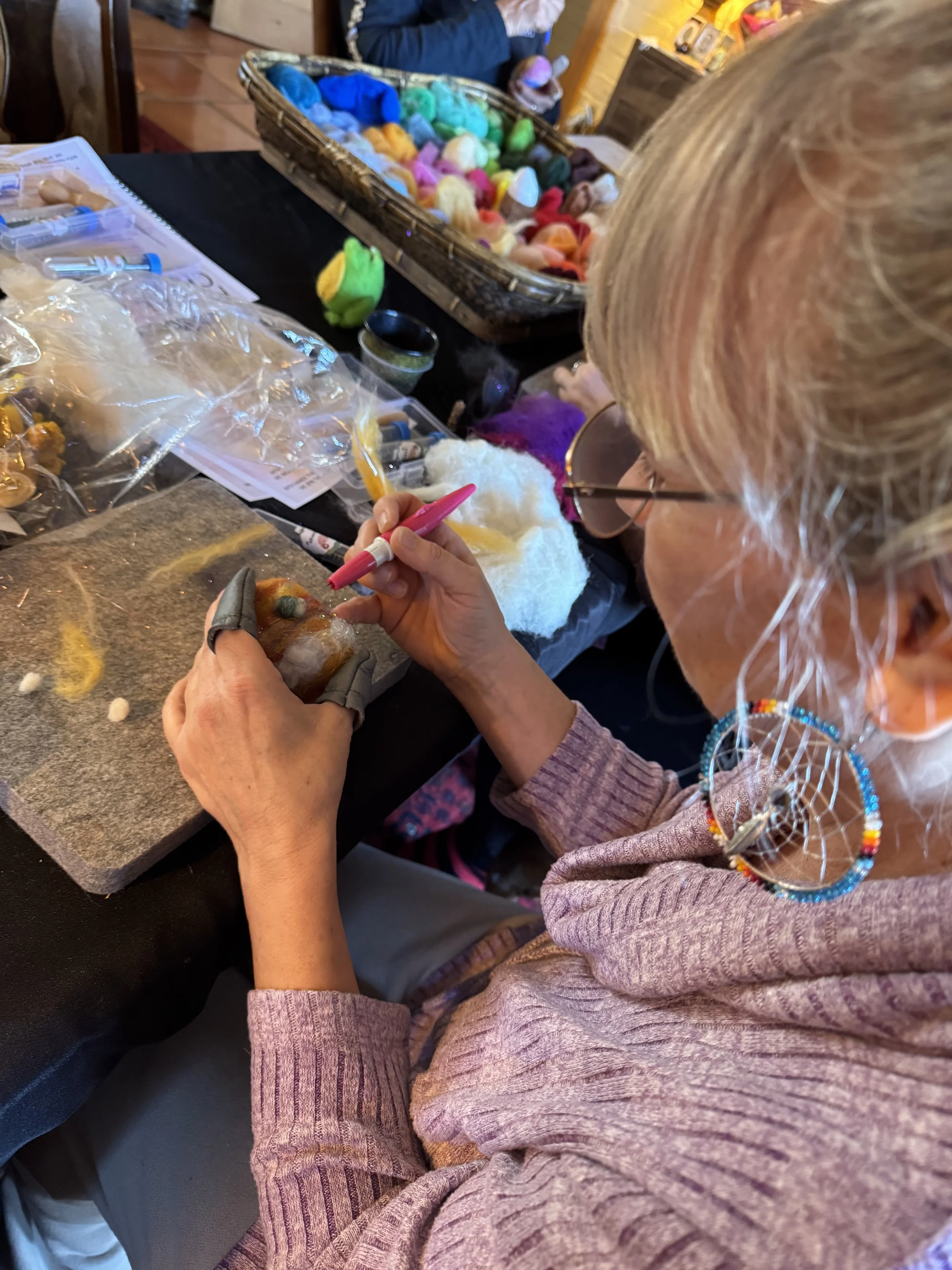 A woman with blonde hair wearing large colorful earrings and glasses is sitting at a table, crafting with wool and felting tools. On the table are various colors of wool, felting supplies, and a basket of multicolored yarns. She is focused on her art