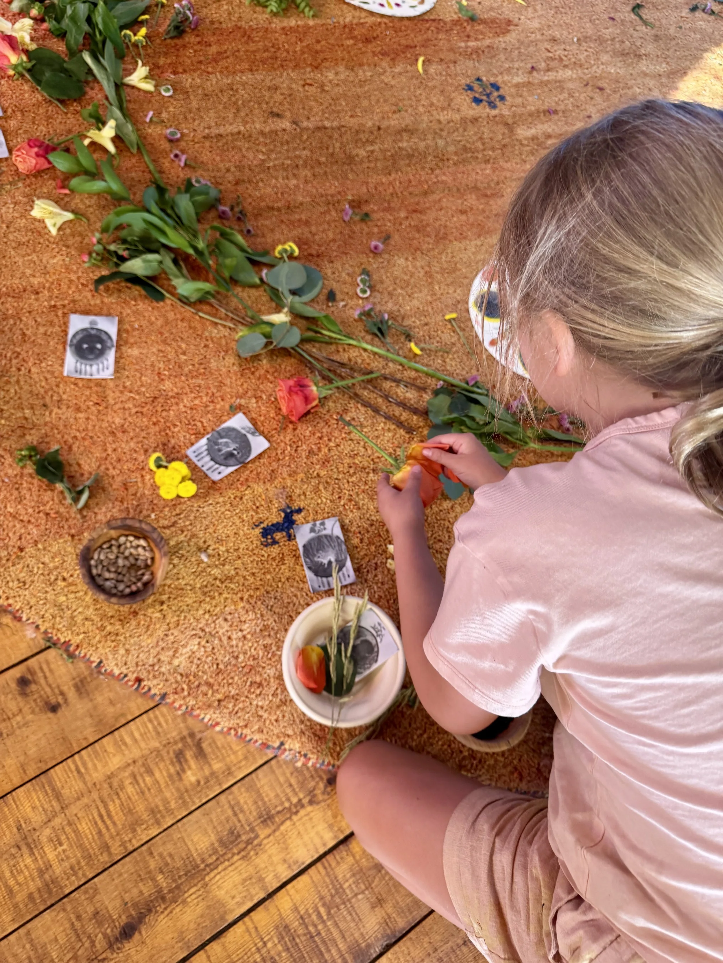 A young girl arranging flowers on a circular, orange, textured rug with some flower photographs and small bowls of seeds and flowers nearby.