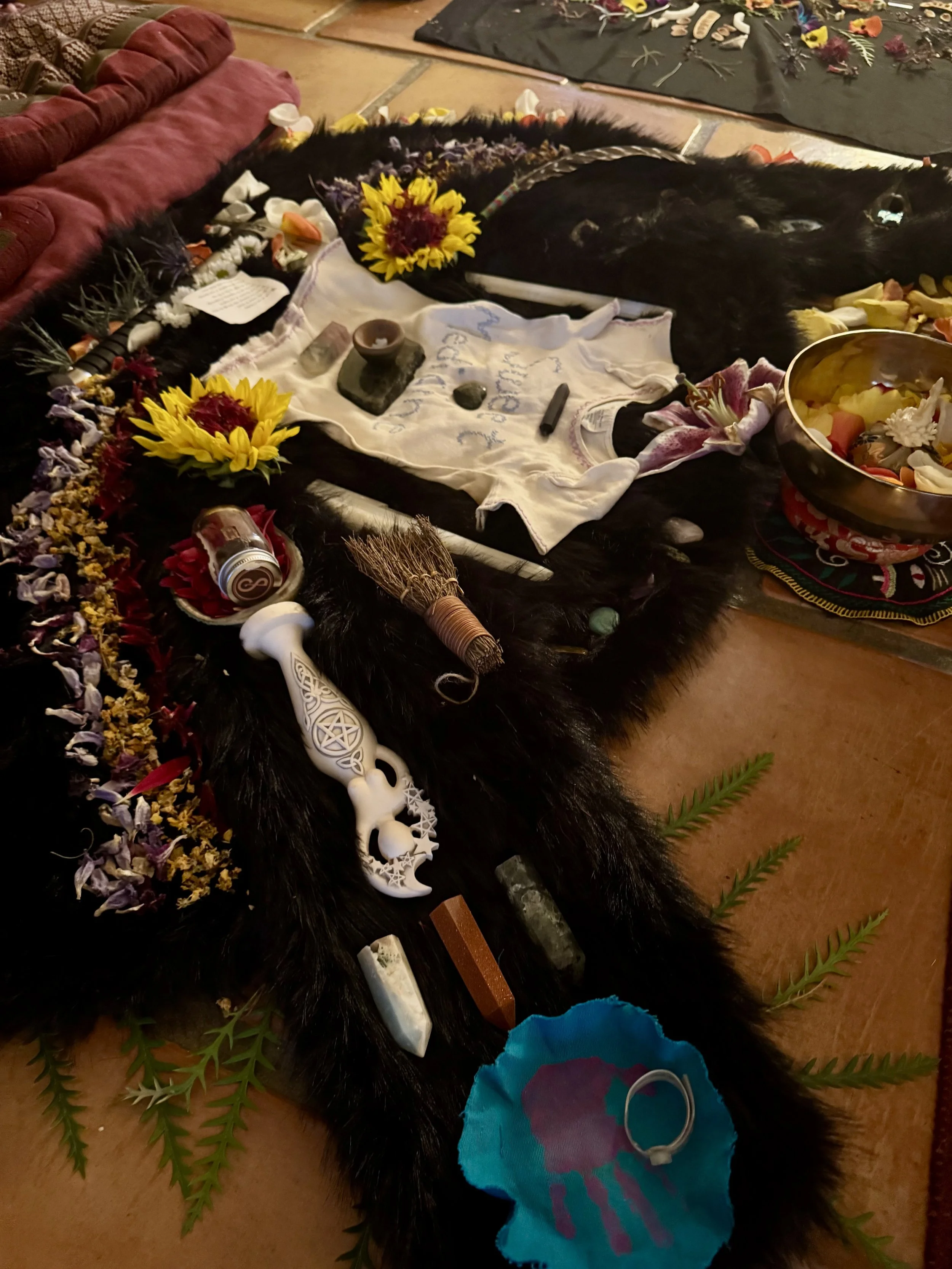 A memorial altar with various items including sunflowers, candles, crystals, dried flowers, a feather, a white decorative dagger, and a ring, arranged on a black fur cloth around a person's body lying down with flowers placed on them, on a tiled floo