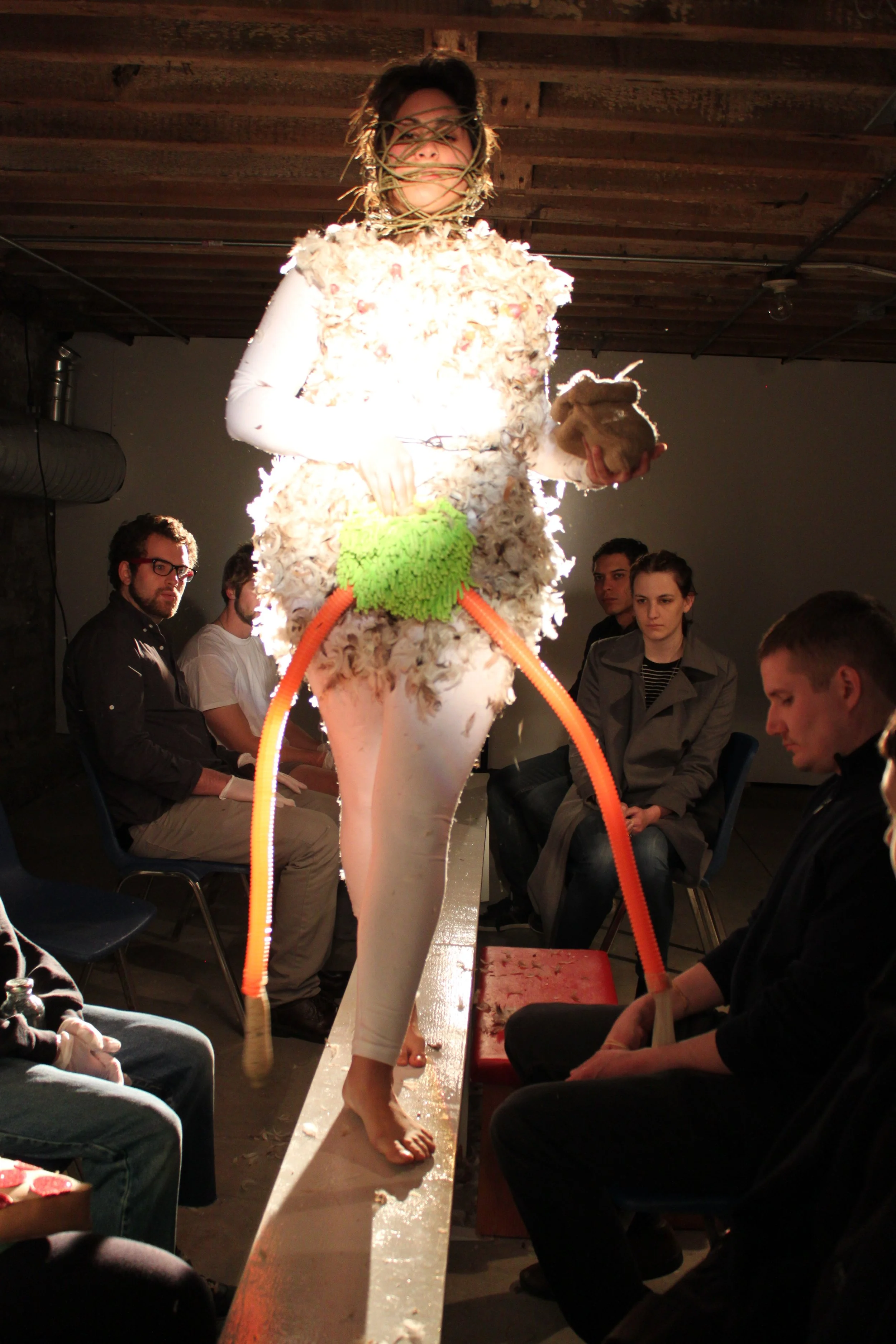 A person walking on a narrow platform wearing an avant-garde costume that resembles a sheep, with woolly texture and large rubber tubes hanging from the waist, in a room with an audience seated on either side watching the runway show.
