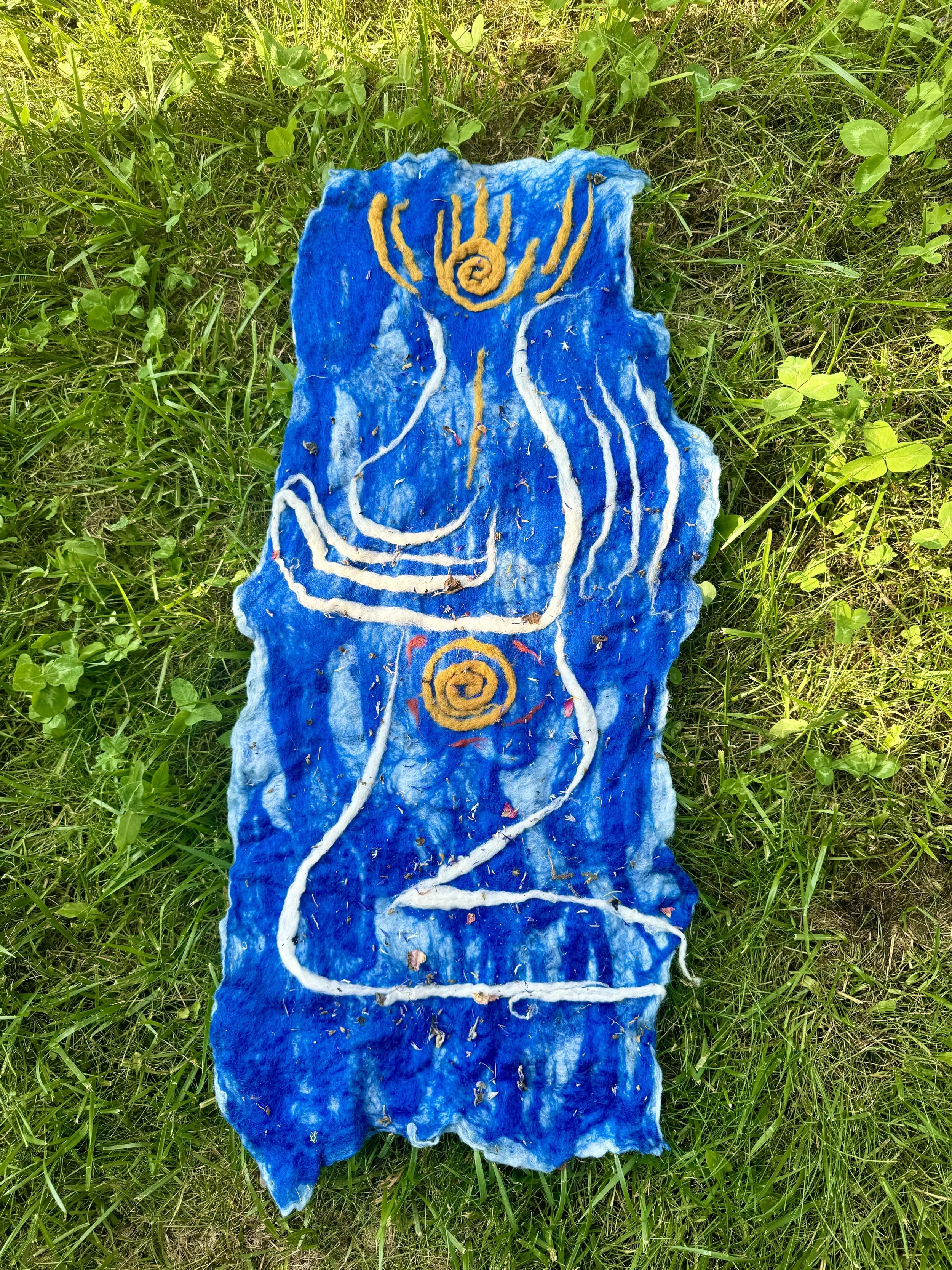 Textile art with abstract human figure on a blue background, featuring yellow and white swirling lines, outdoors on grass.