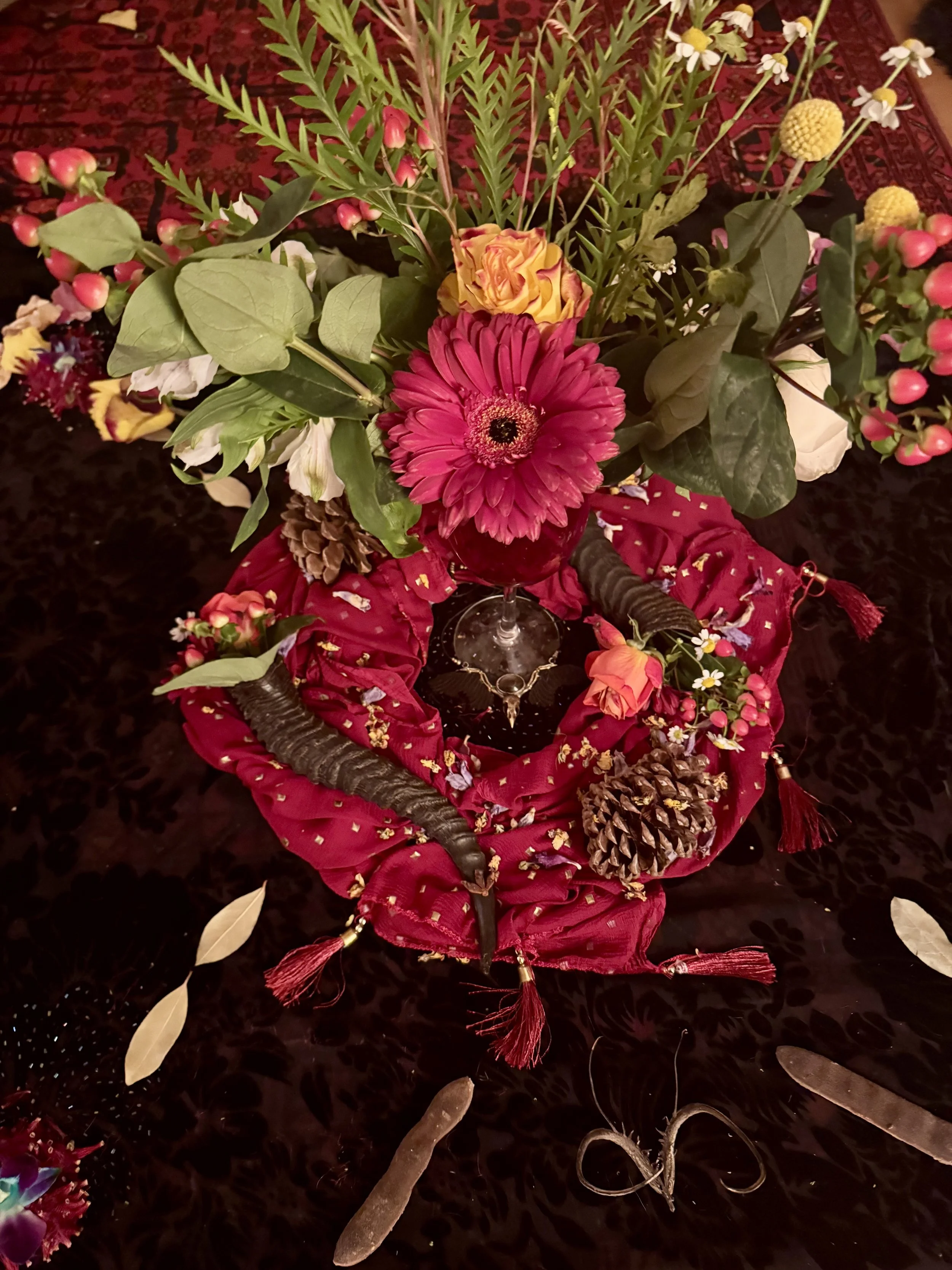 A flower arrangement with various colorful flowers and green leaves, placed on a red fabric and black tablecloth with tassels and pinecones.