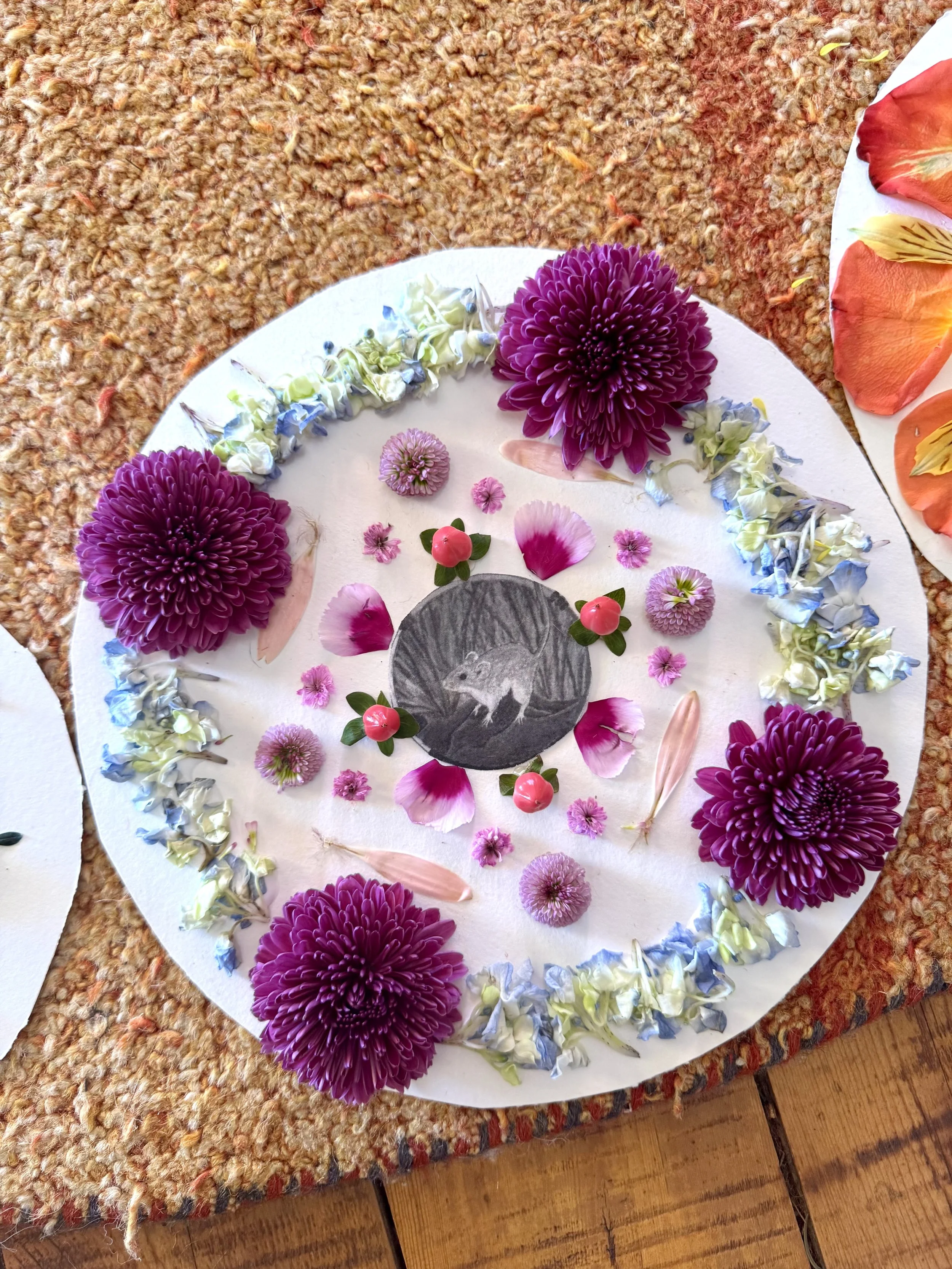 Decorative circular arrangement with purple chrysanthemums, pink and white flower petals, small pink berries with green leaves, and a black and white drawing of a squirrel in the center, on a white circular cloth. The arrangement is on a textured ora