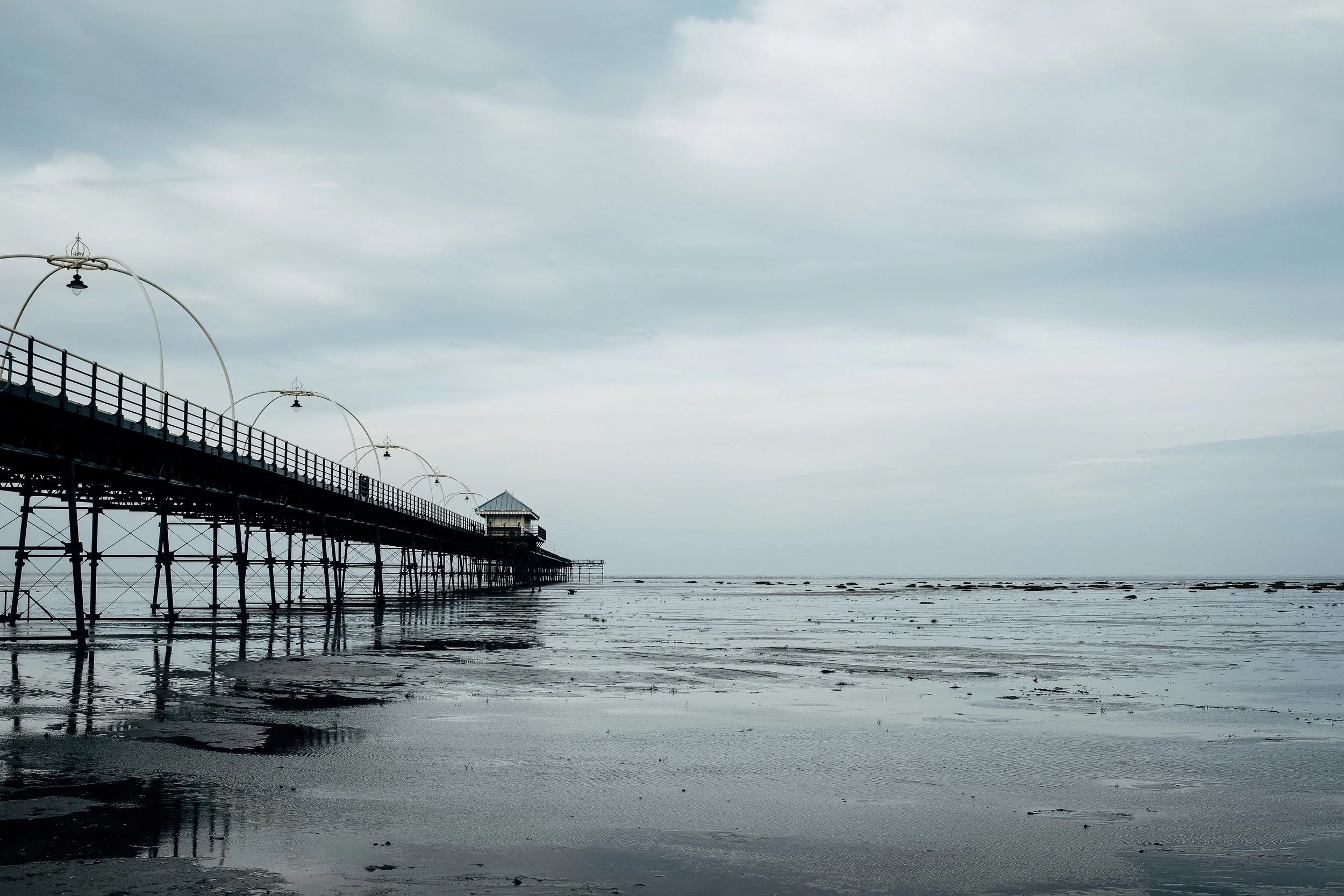 A long pier extends over the wet sand in a beach, with a small building at the end and street lamps along its length. The sky is cloudy and the water is calm.
