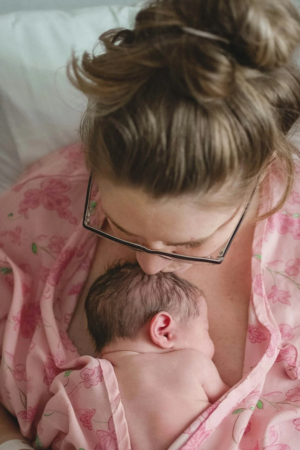 A woman with glasses gently holding a newborn baby on her chest, the baby is sleeping wrapped in a pink blanket with floral patterns.