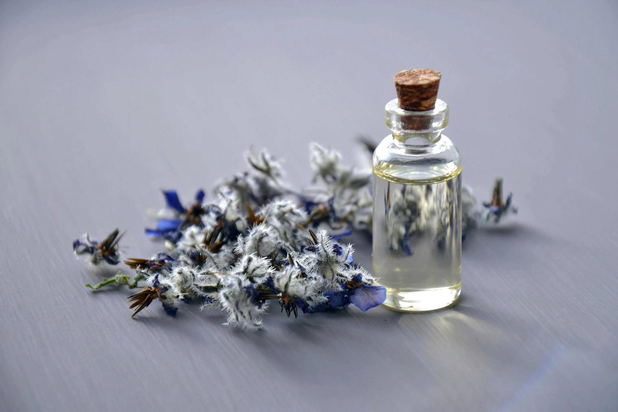 Une petite bouteille en verre avec un bouchon en liège, contenant une huile claire, posée à côté d'un bouquet de fleurs séchées bleues, blanches et violettes sur une surface gris clair.
