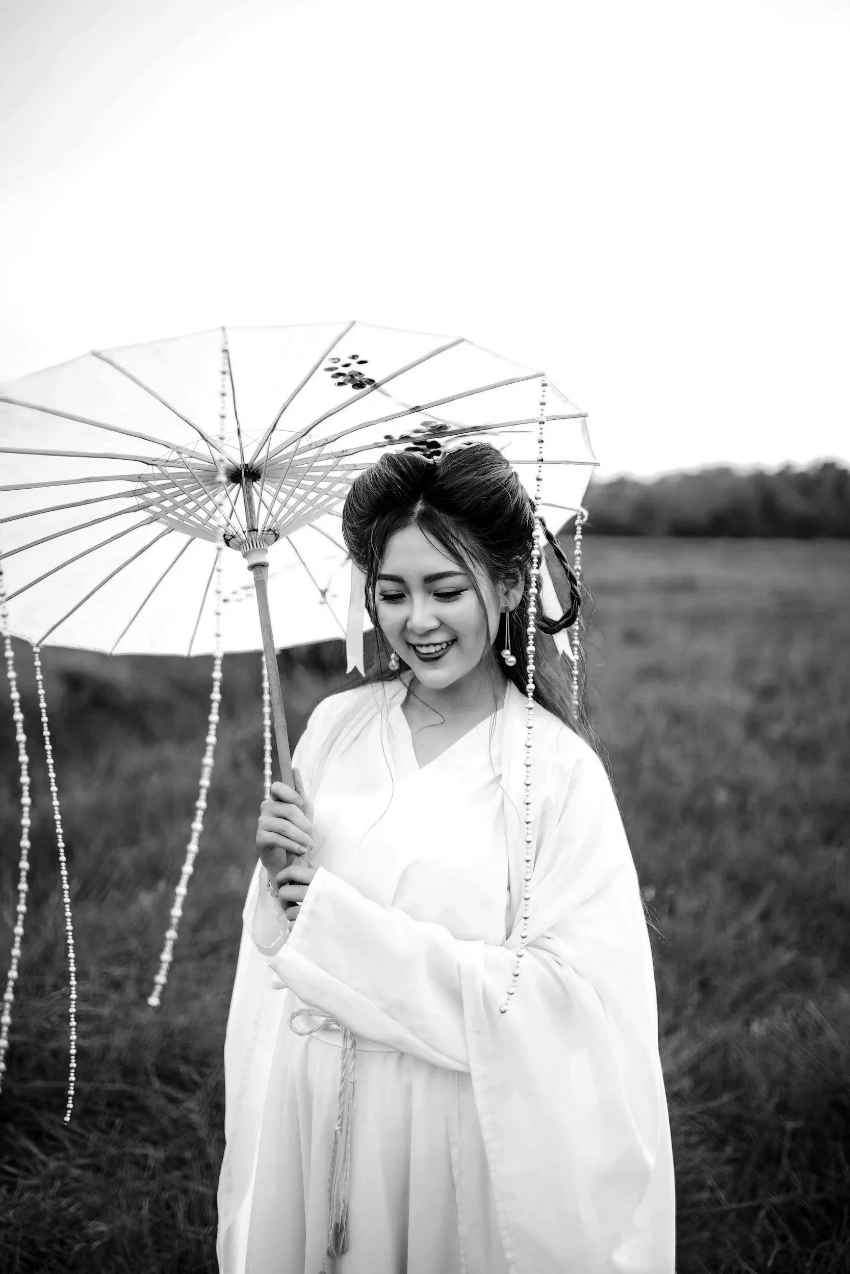 Une femme en kimono traditionnel japonais avec un parapluie en papier dans un champ, souriant en regardant vers le bas, dans un style noir et blanc.