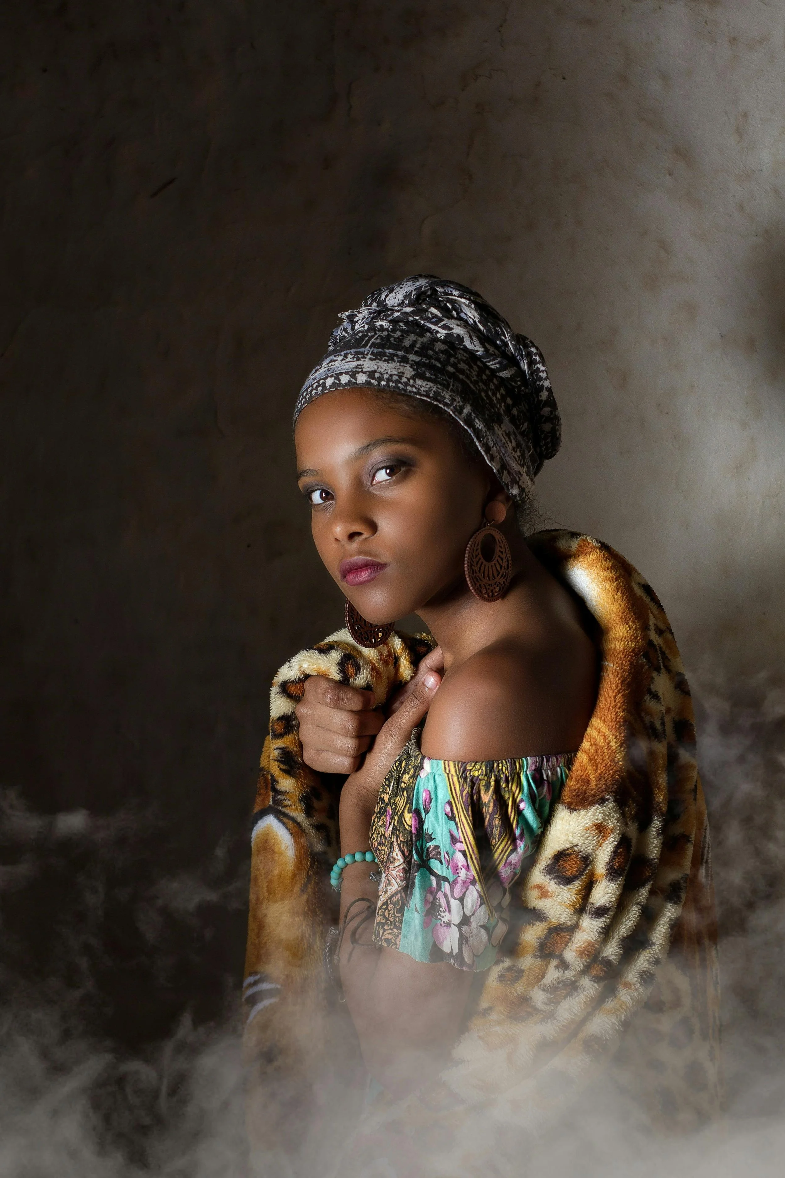 Jeune femme africaine portant une robe colorée, des boucles d'oreilles en bois, un foulard imprimé et une veste en fausse fourrure léopard, dans un décor sombre avec de la brume au premier plan.