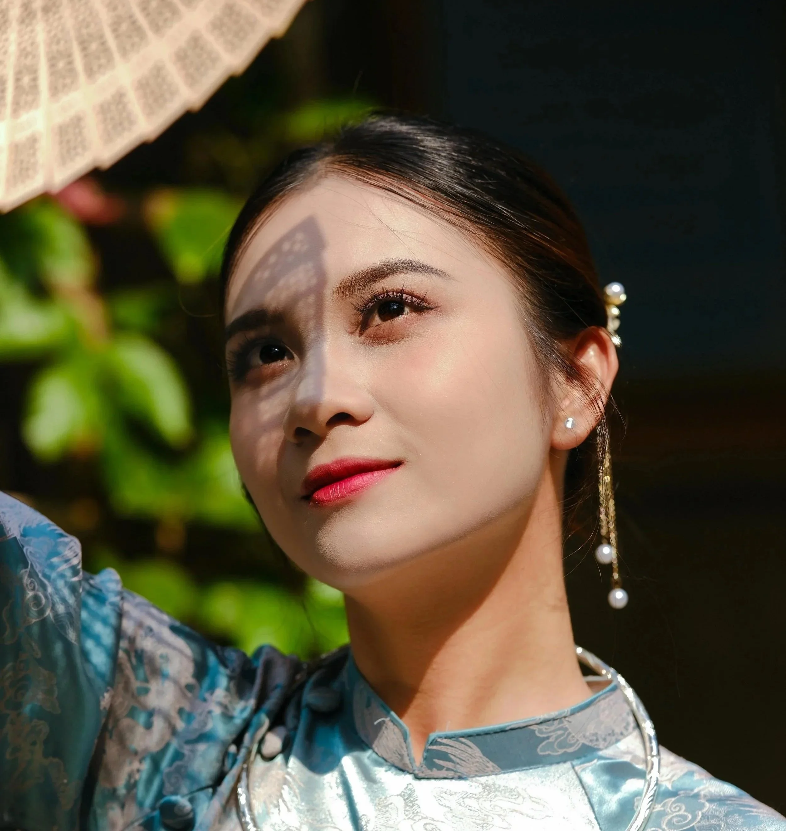 Portrait d'une femme asiatique en tenue traditionnelle asiatique bleue, regardant vers le haut avec un parasol en arrière-plan, soleil brillant sur son visage.