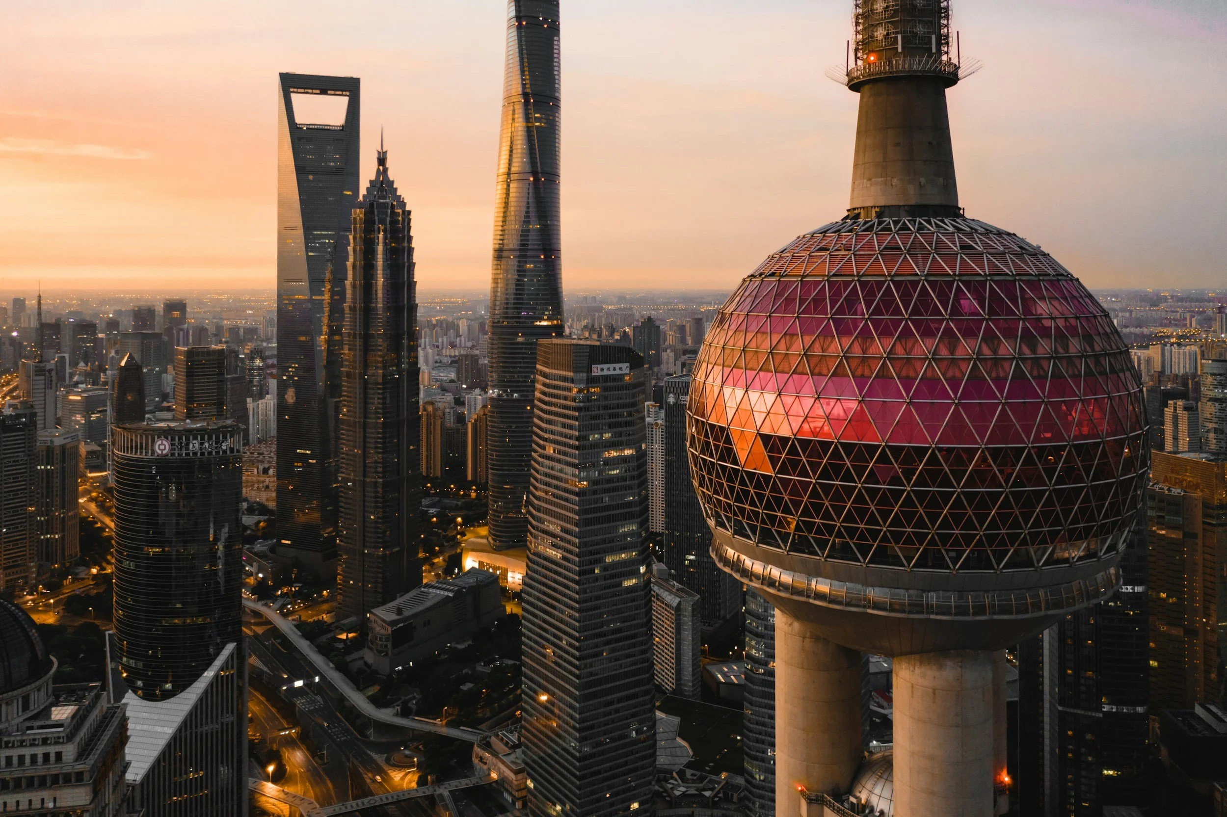 A city skyline at sunset featuring tall skyscrapers, including one with a spherical observation deck reflecting pink and orange hues, in a modern metropolis, possibly Shanghai, China.