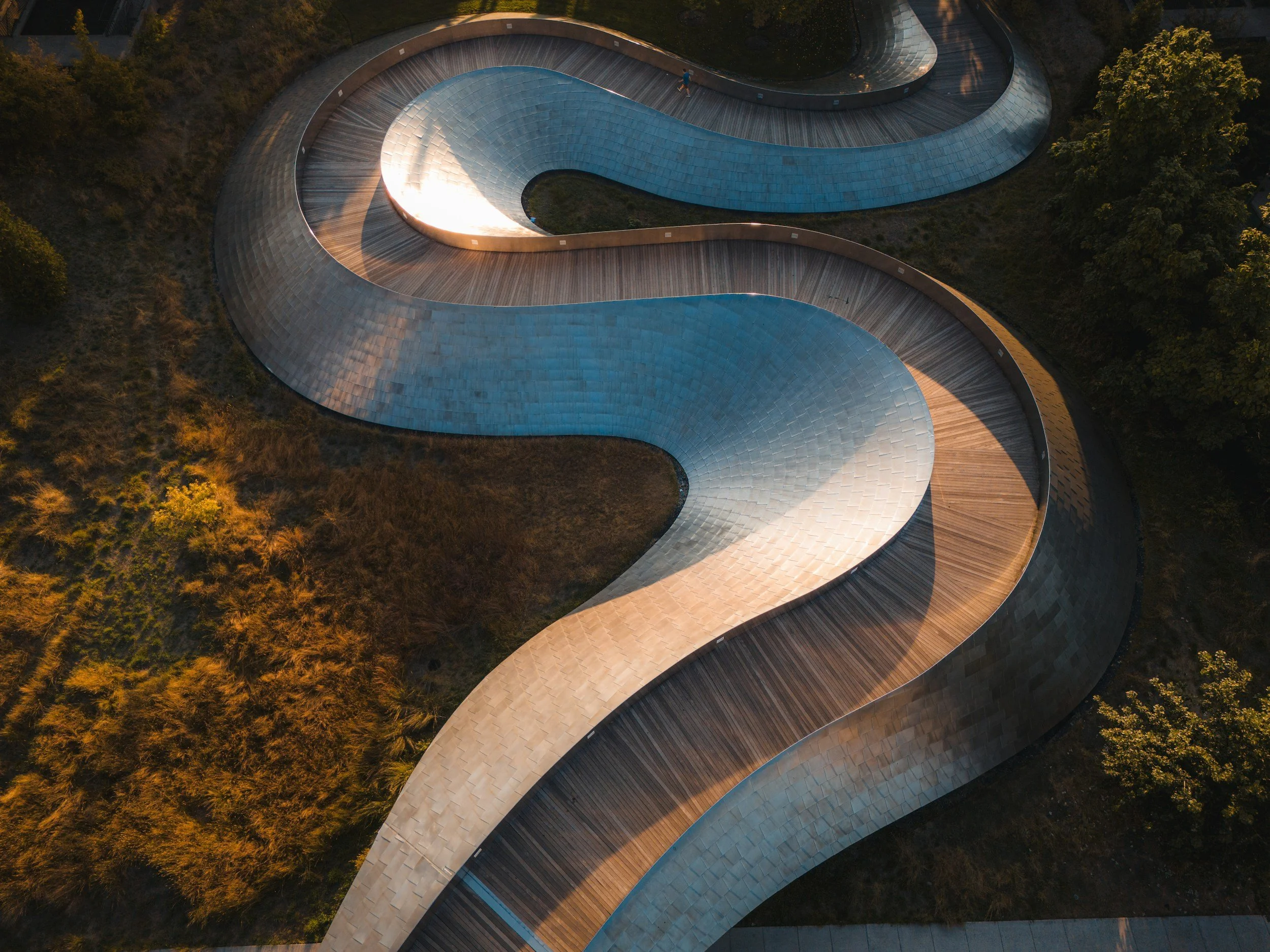 Aerial view of a winding, modern pedestrian bridge with metallic surface and wooden walkway, surrounded by trees and grass.