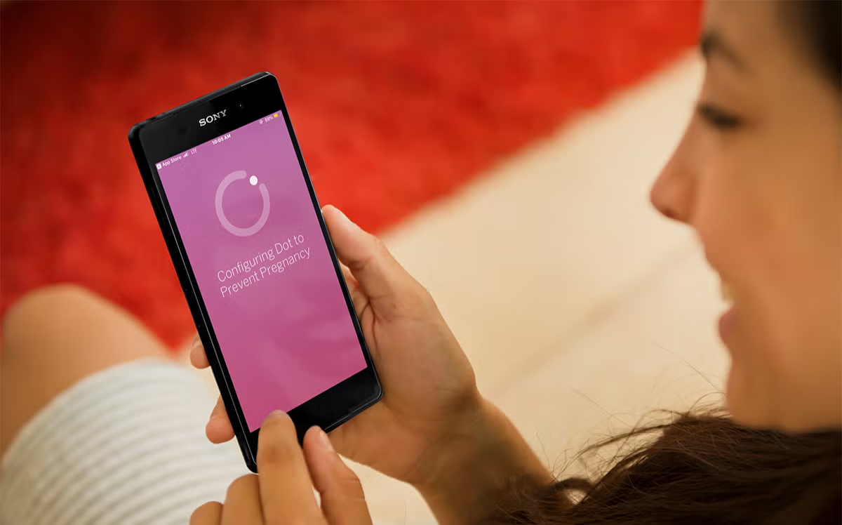 A woman smiling while holding a smartphone that displays an app configuring a dot to prevent pregnancy.