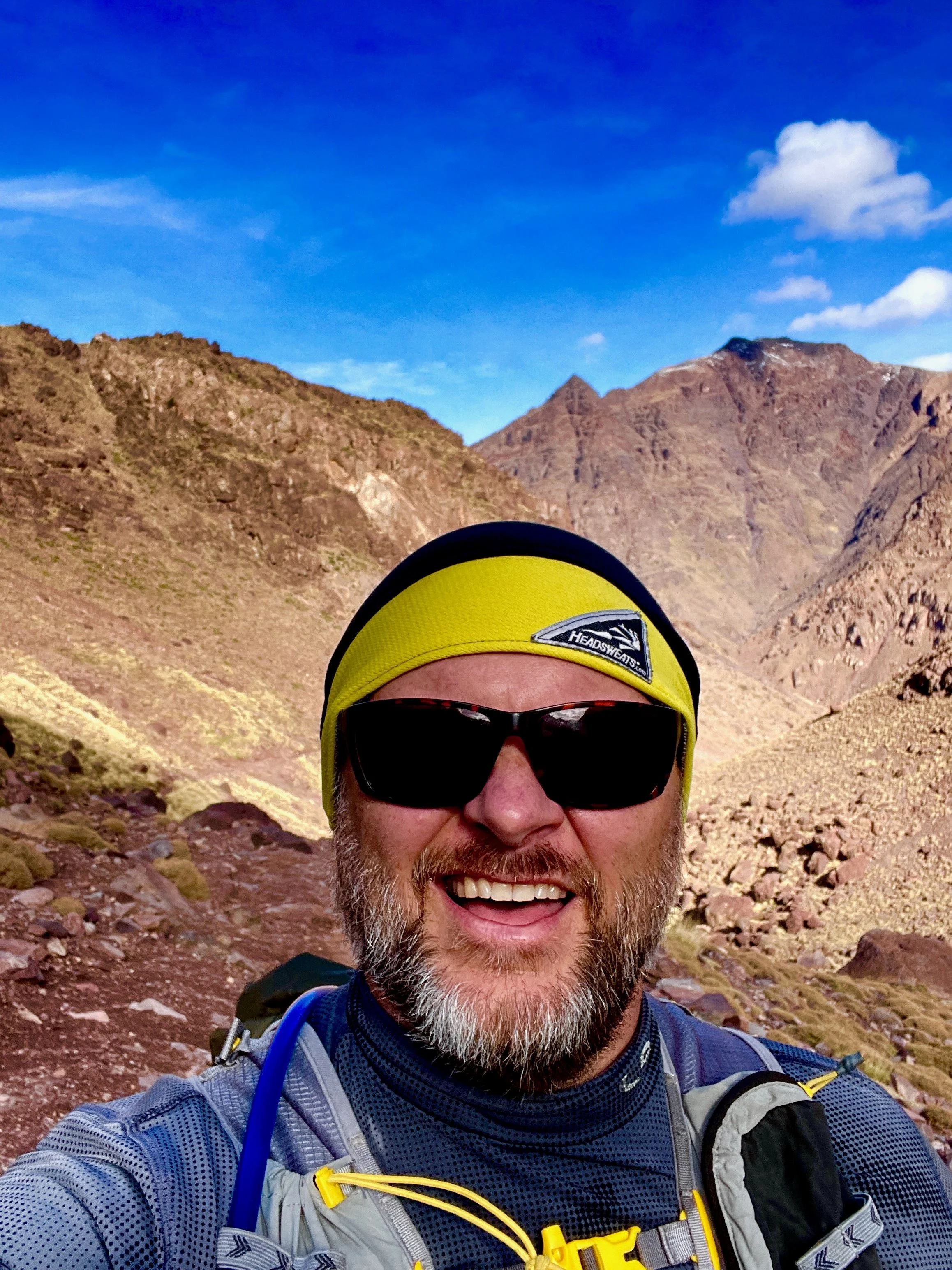A man with a beard wearing a yellow headband, sunglasses, and outdoor gear, smiling with mountains and a blue sky in the background.
