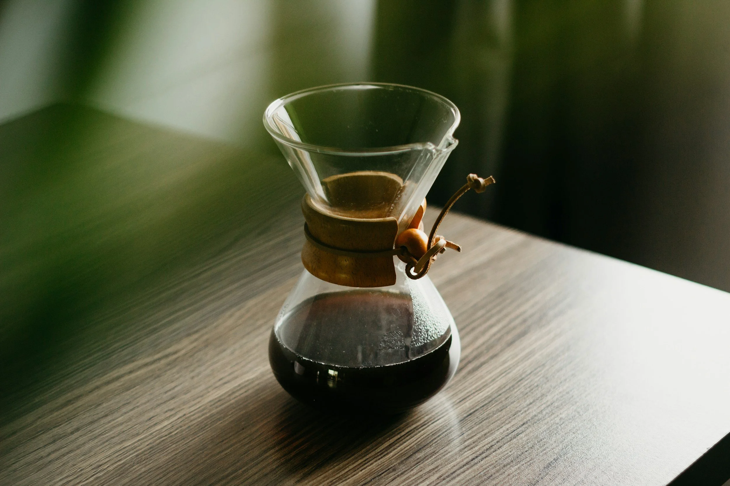 A glass pour-over coffee carafe with dark coffee inside, sitting on a wooden surface with a blurred green background.