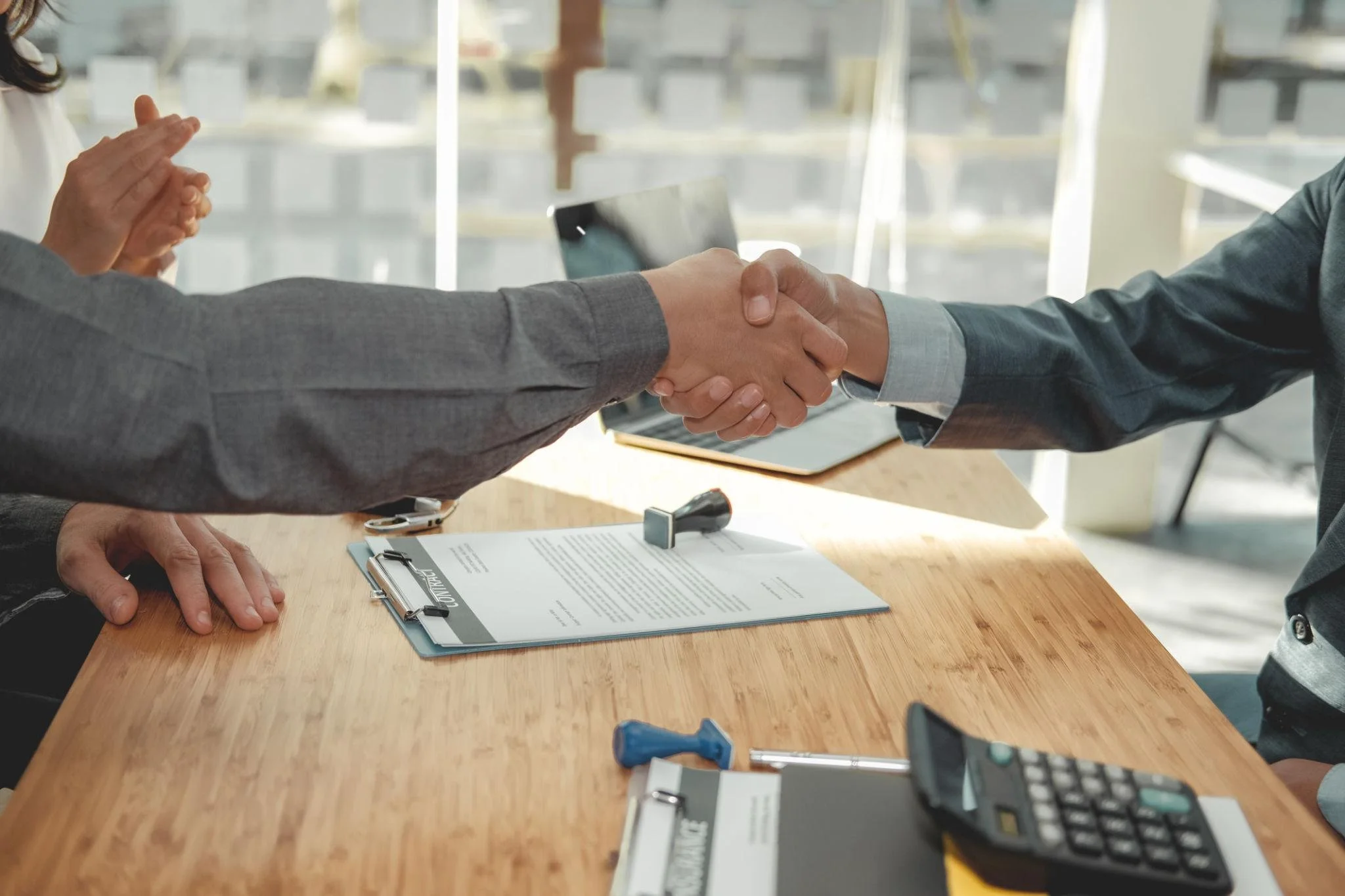 Two people shaking hands over a desk in what appears to be a business meeting or interview, with documents, a calculator, and office supplies on the table.