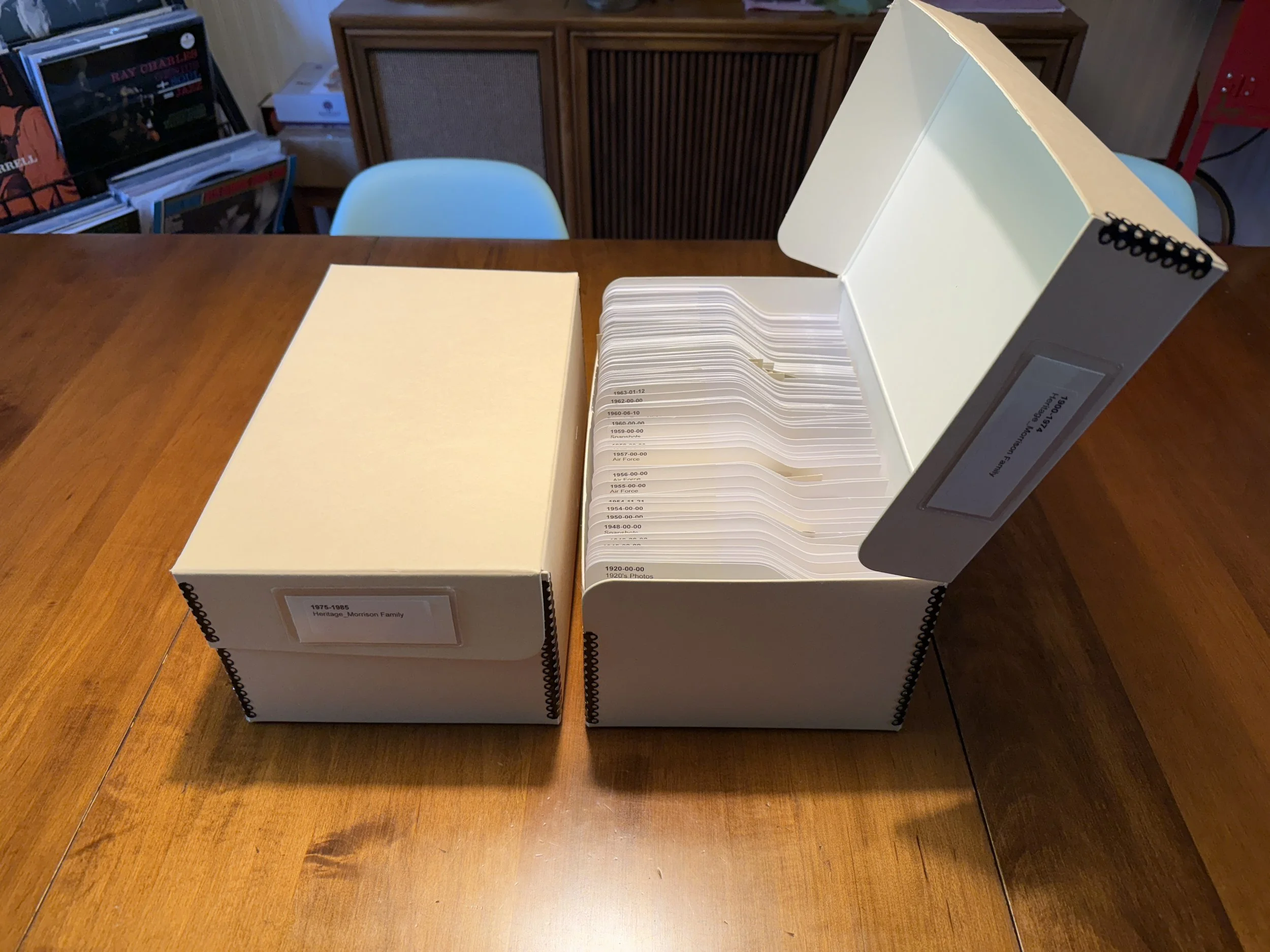 A archival storage box containing organized printed photos on a wooden table, with a label indicating it contains heritage family photos from 1975 to 1985, and a side section showing year labels from 1920 to 1965.