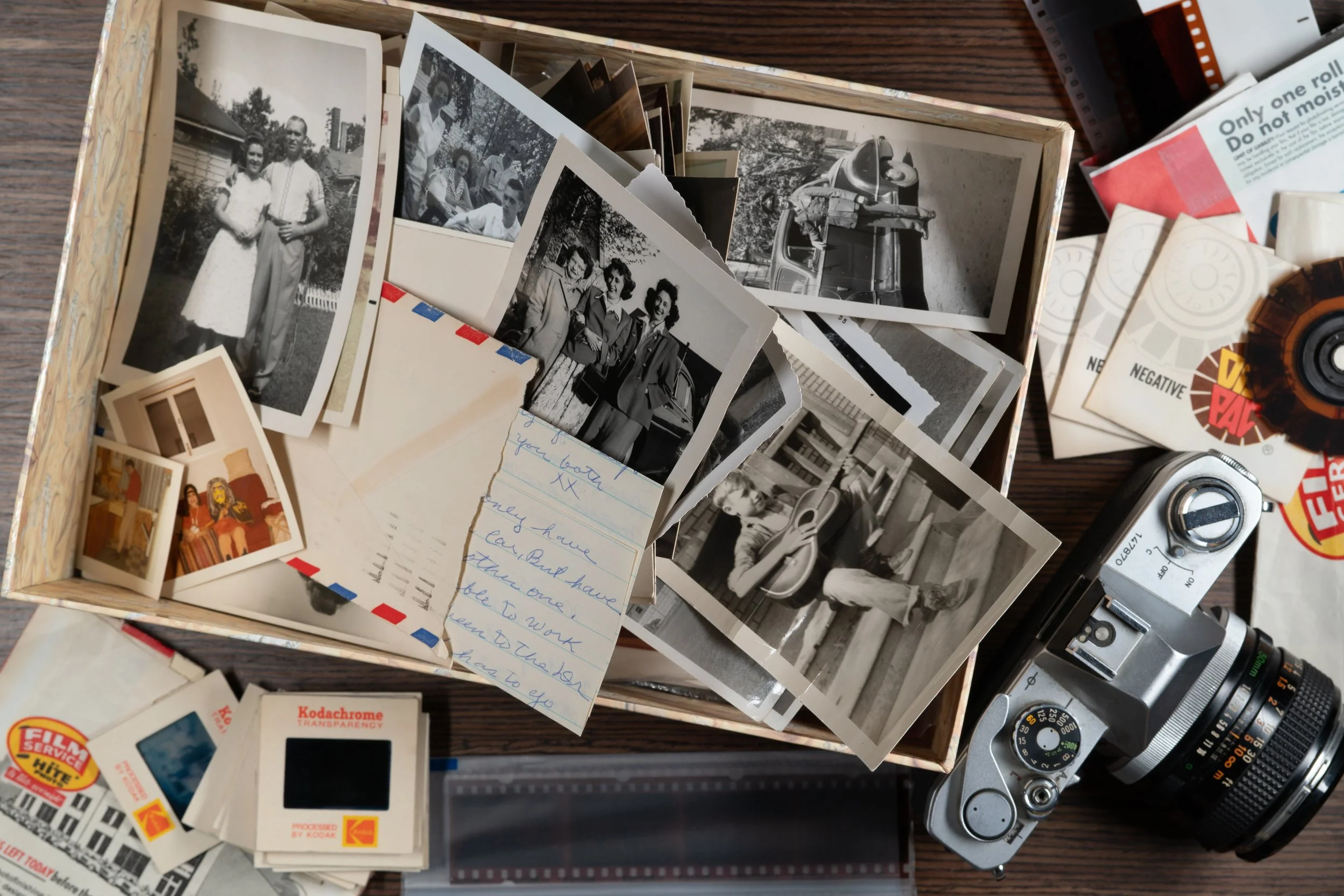 A collection of black and white photographs, vintage film photographs, a handwritten note, film reels, and camera on a wooden surface that need organizing