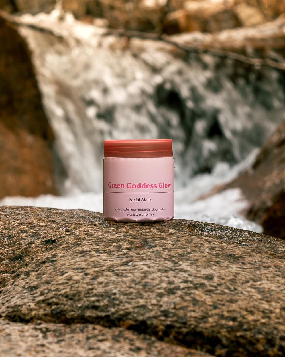 Green Goddess Glow facial mask jar photographed on a rock beside a flowing waterfall, featuring natural skincare packaging in an outdoor nature setting.