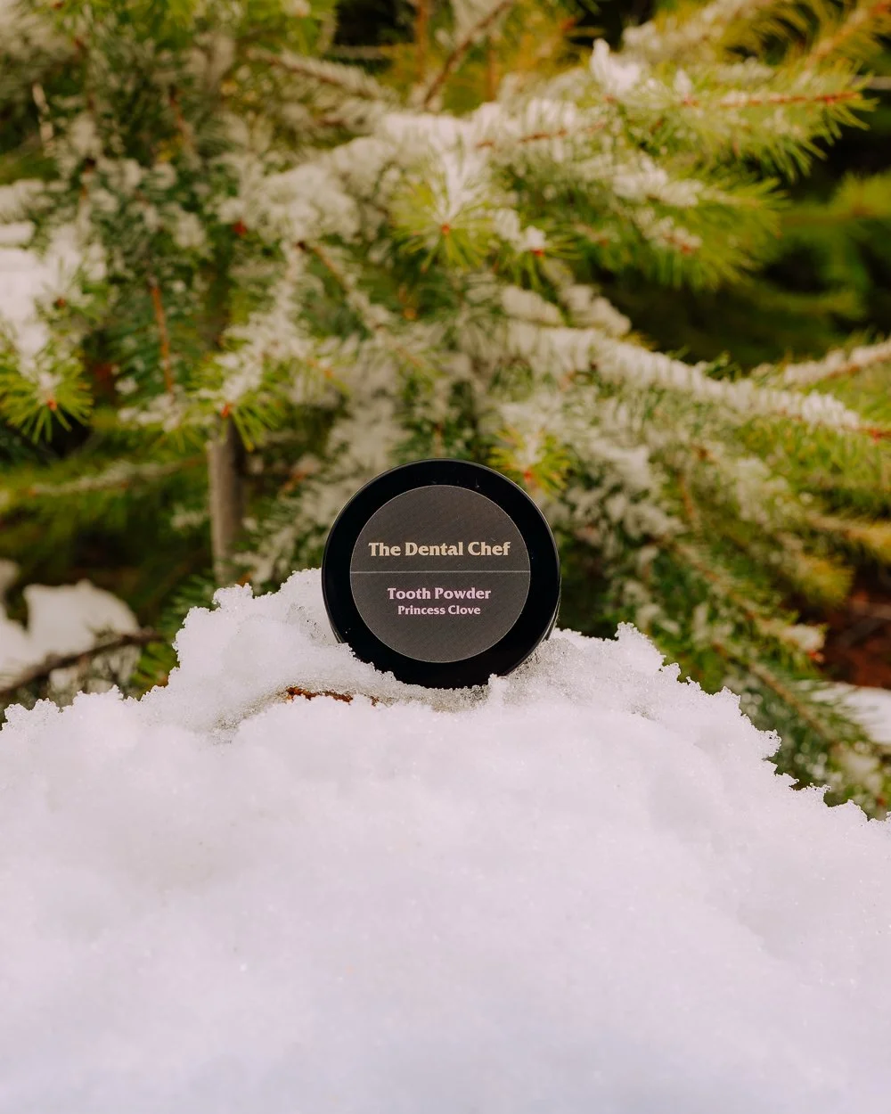 The Dental Chef Princess Clove tooth powder jar photographed outdoors on fresh snow with evergreen trees in the background, showcasing natural oral care packaging in a winter setting.