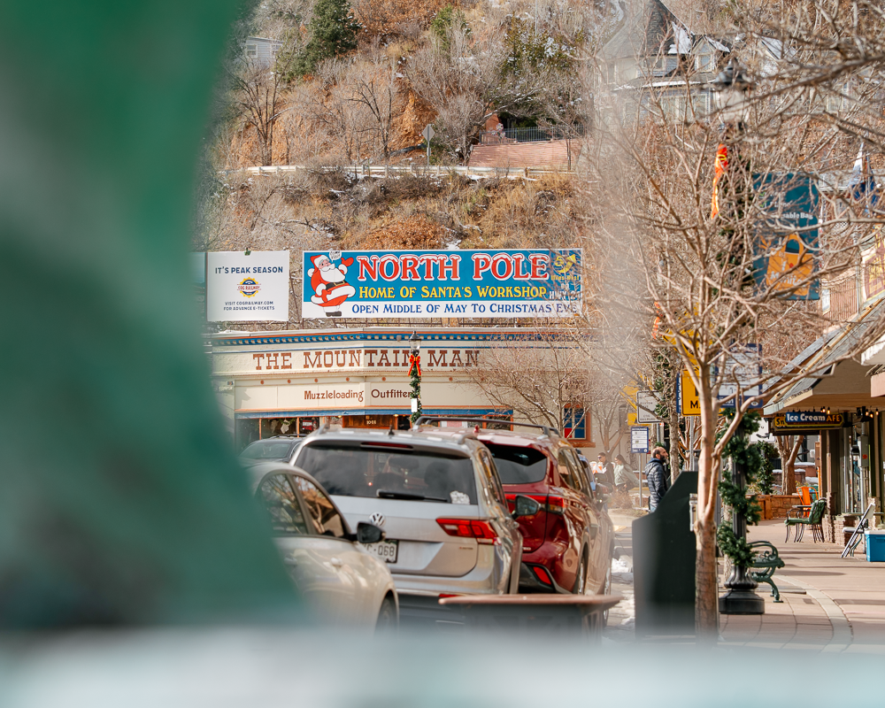Creative telephoto shot of the North Pole Santa’s Workshop sign in Manitou Springs, framed through a blurred foreground with mountain scenery.