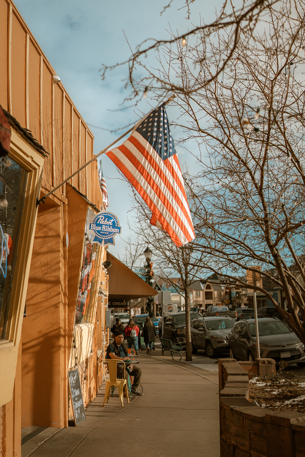 Warm street photo in Manitou Springs showing an American flag, local storefronts, and people enjoying the downtown atmosphere.